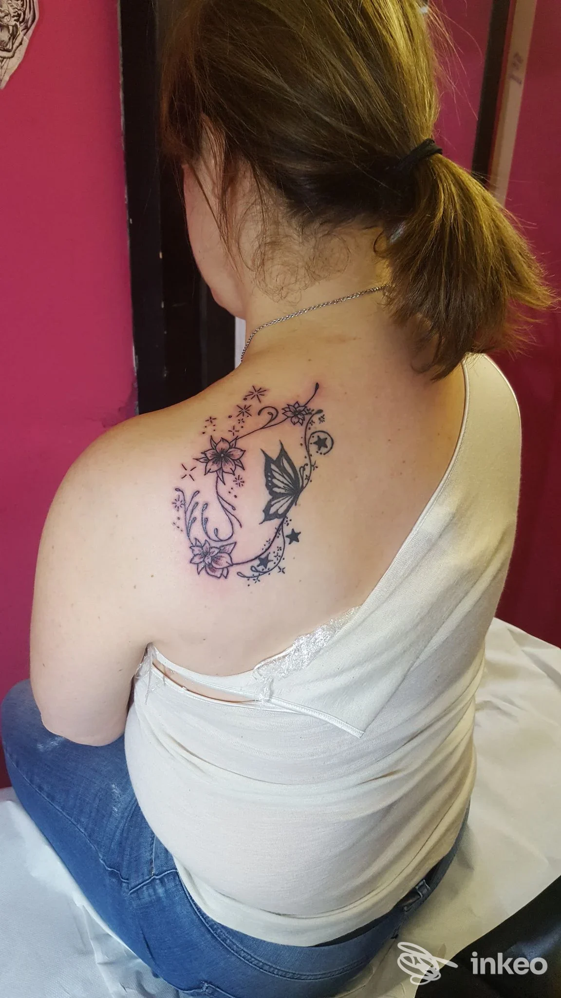 Tattoo floreale stilizzato visibile sulla spalla destra della persona vista di schiena nell’ambiente di uno studio di tatuaggi a Piacenza.