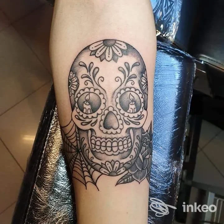 Tatuaggio stile sugar skull sul braccio, disegno in ambito ornamentale con contorni neri e motivi floreali; nulla di visibile sul volto.