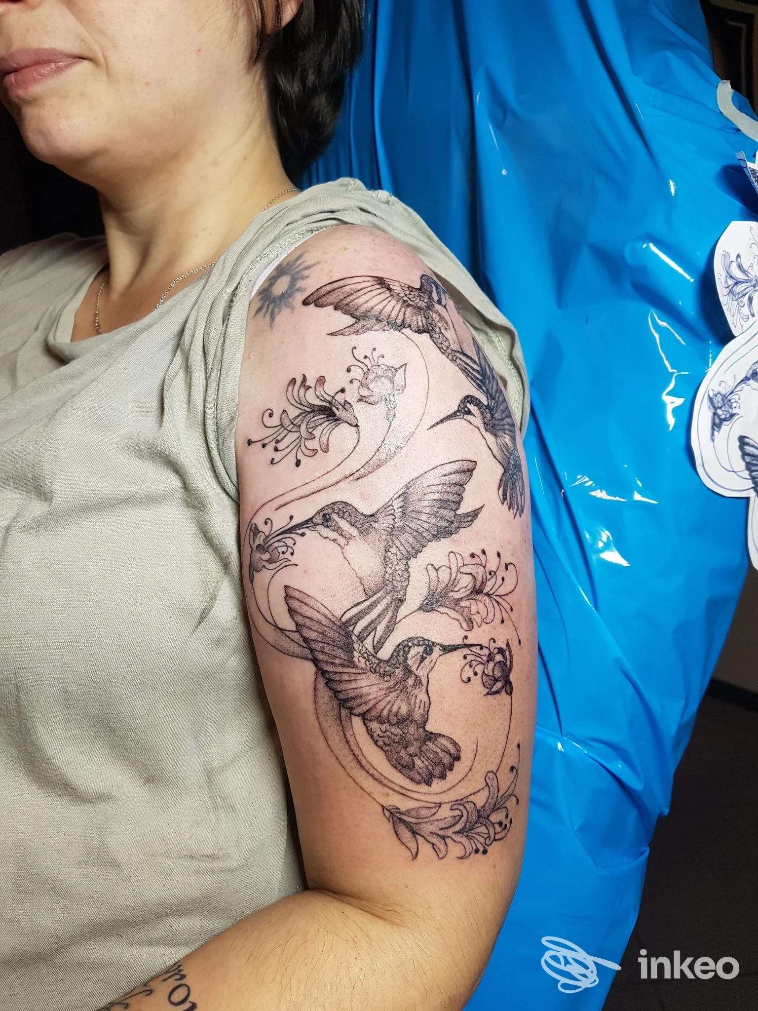 Tattoo a braccio superiore visibile: uccelli in volo tra motivi floreali su pelle chiara, su sfondo ambiente studio con plastica blu. Studio Laura Capellini Tattoo Studio, Salsomaggiore Terme.