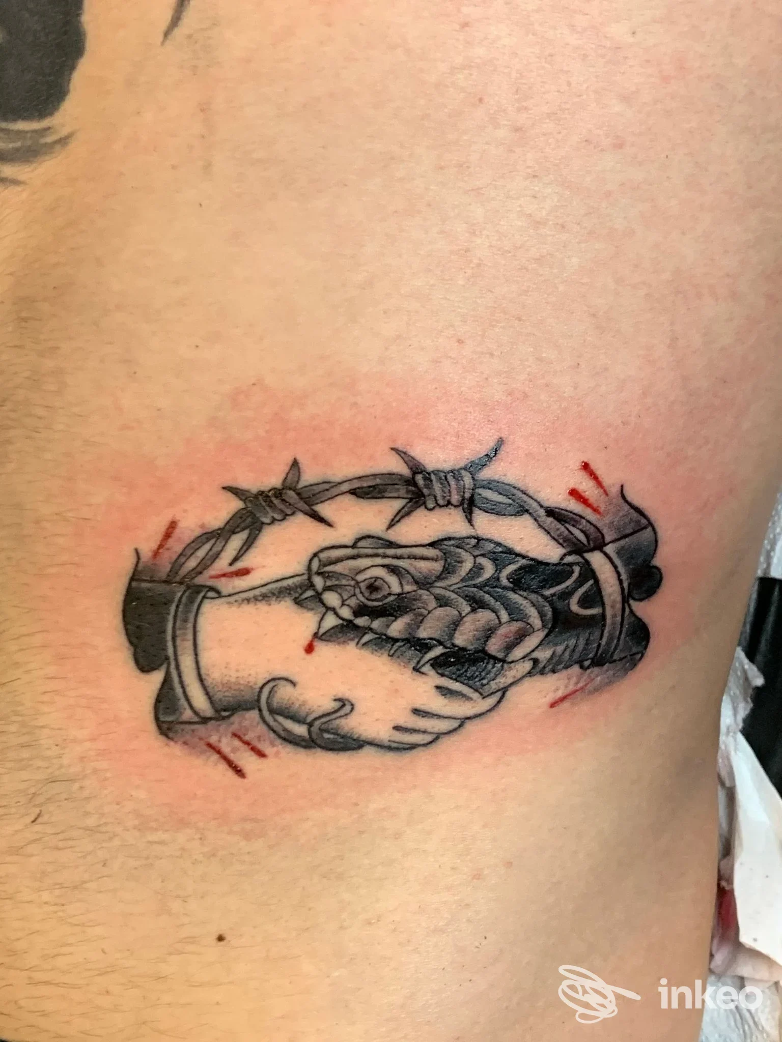 Tattoo su avambraccio raffigurante due mani che si stringono, circondate da filo spinato, con riflessi rossi; pelle visibile e contesto privo di testo o elementi di social media.
