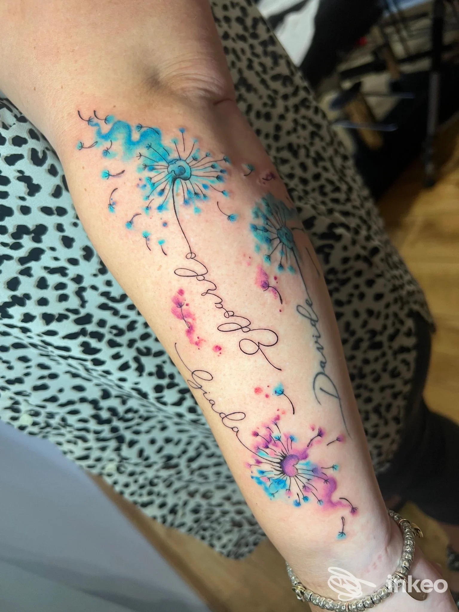 Tattoo floreali a gel d’acqua sull’avambraccio, con dente di dandelion stilizzato in toni azzurri e rosa, firma elegante lungo la pelle. Studio Velvet Tattoo a Forlì.