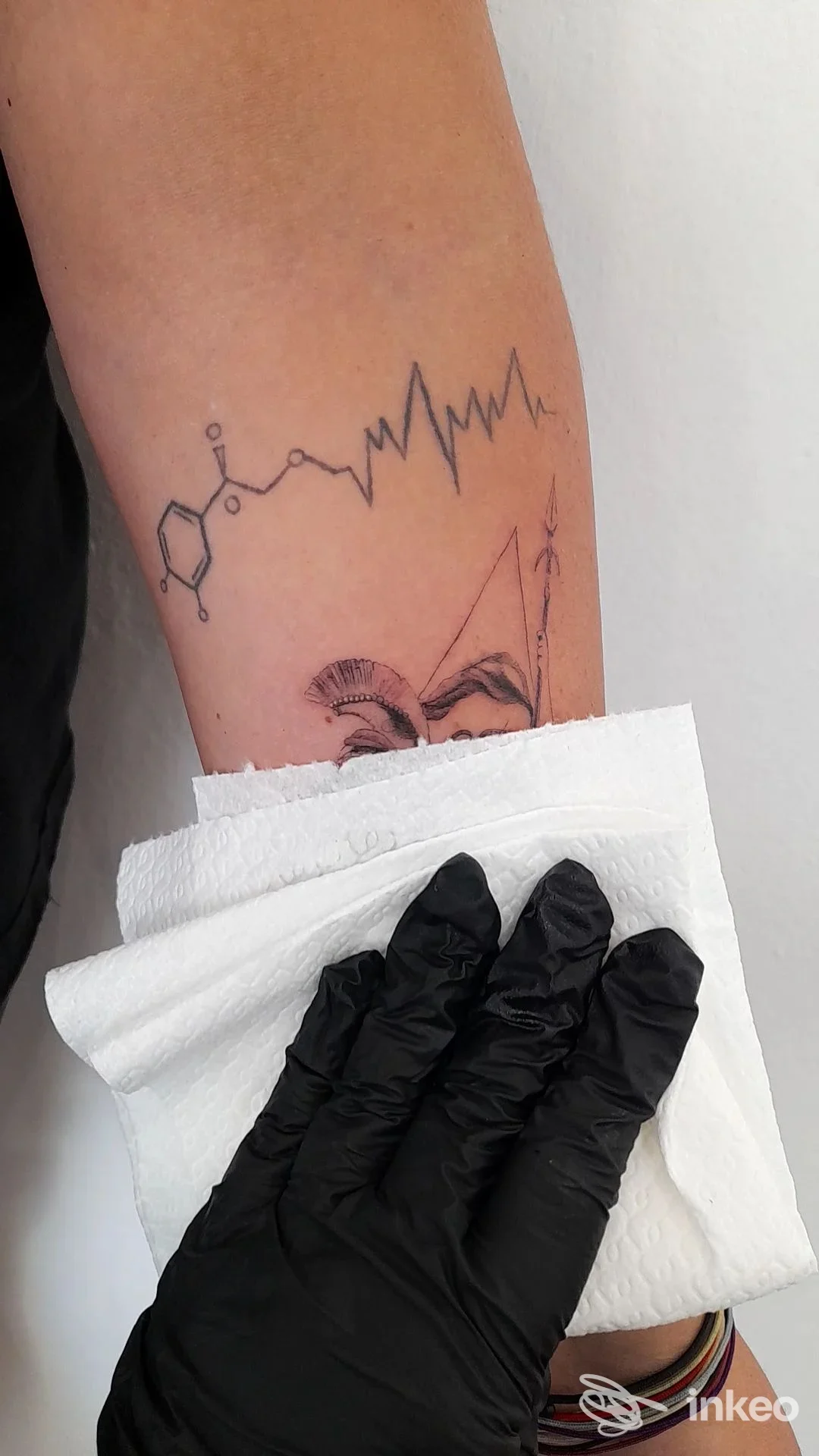 Tattoo sull’avambraccio: linea a onde simile a un battito cardiaco che si estende lungo la pelle, con una piccola grafica chimica. mano guantata e asciugamano bianche visibili.
