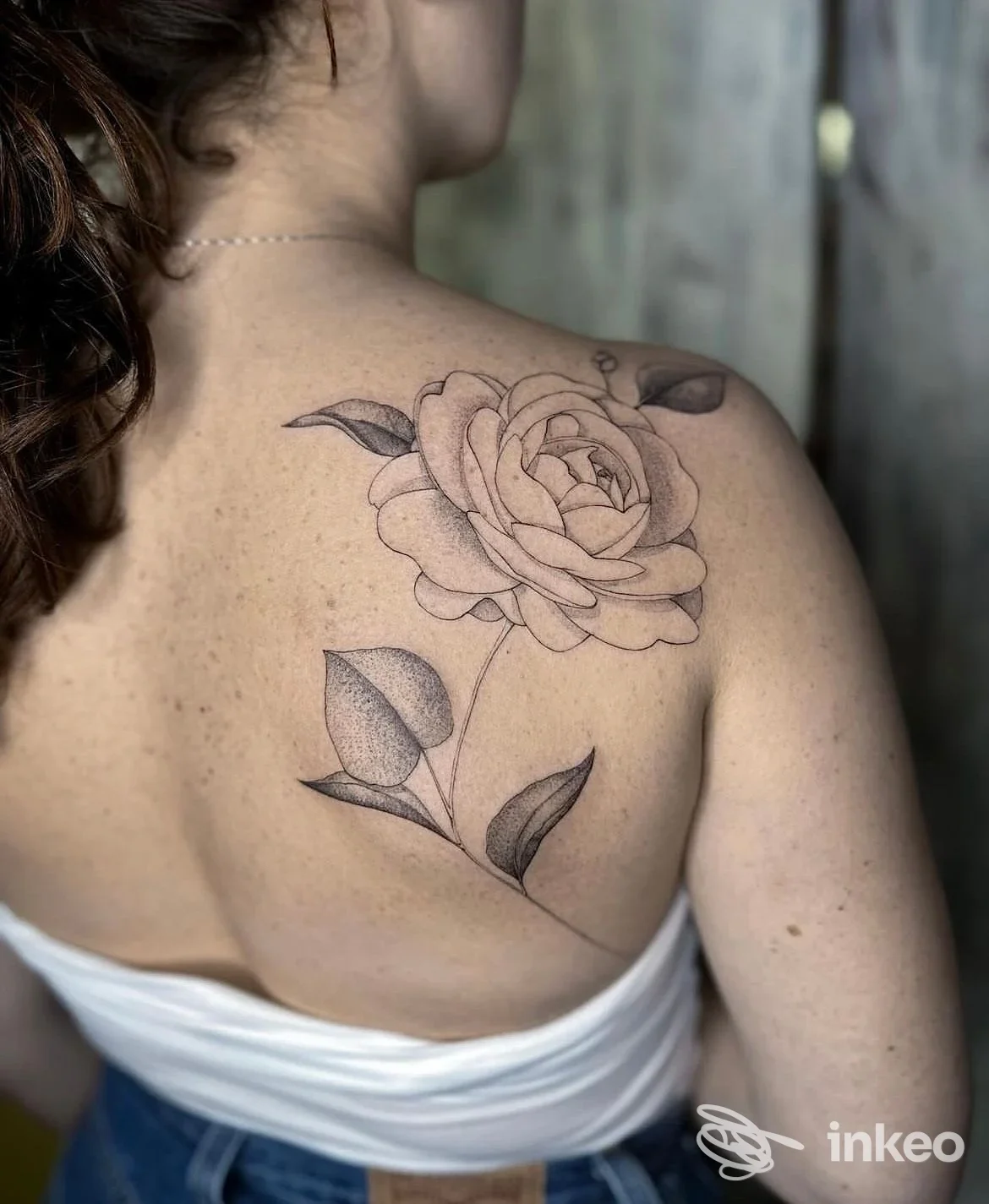Tattoo reale su pelle: grande rosa ben definita sulla spalla e sulla parte superiore della schiena di una donna, con foglie sottostanti.