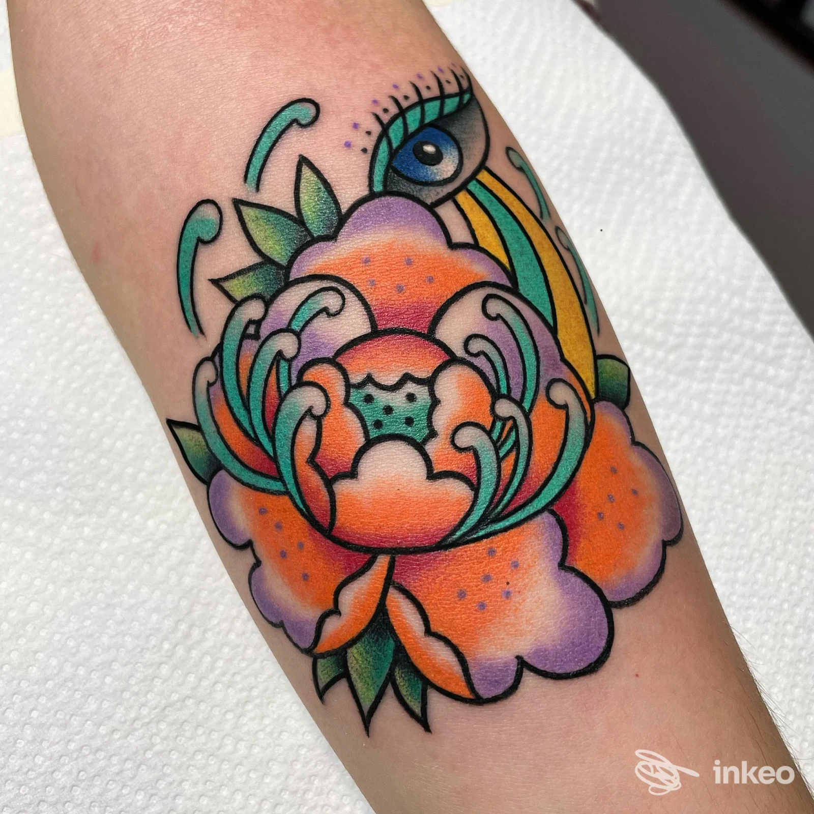 Tattoo colorato su avambraccio raffigurante un grande fiore stilizzato con sfumature arancione, rosa e viola, bordi neri netti, foglie verdi, visibile in una galleria di pelle reale; studio Underskintattoo, Bologna.