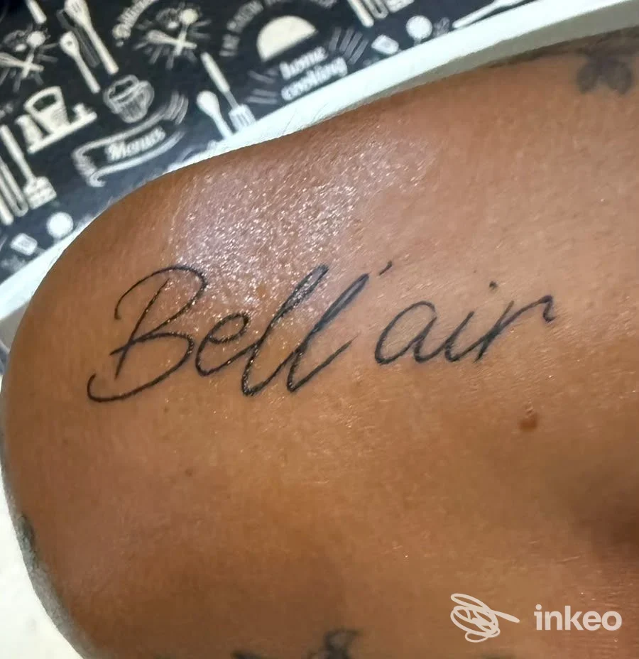 Tattoo in corsivo con la parola Bell'air sulla pelle di una persona, visibile su spalla/brazo, sfondo con pattern nero e bianco.