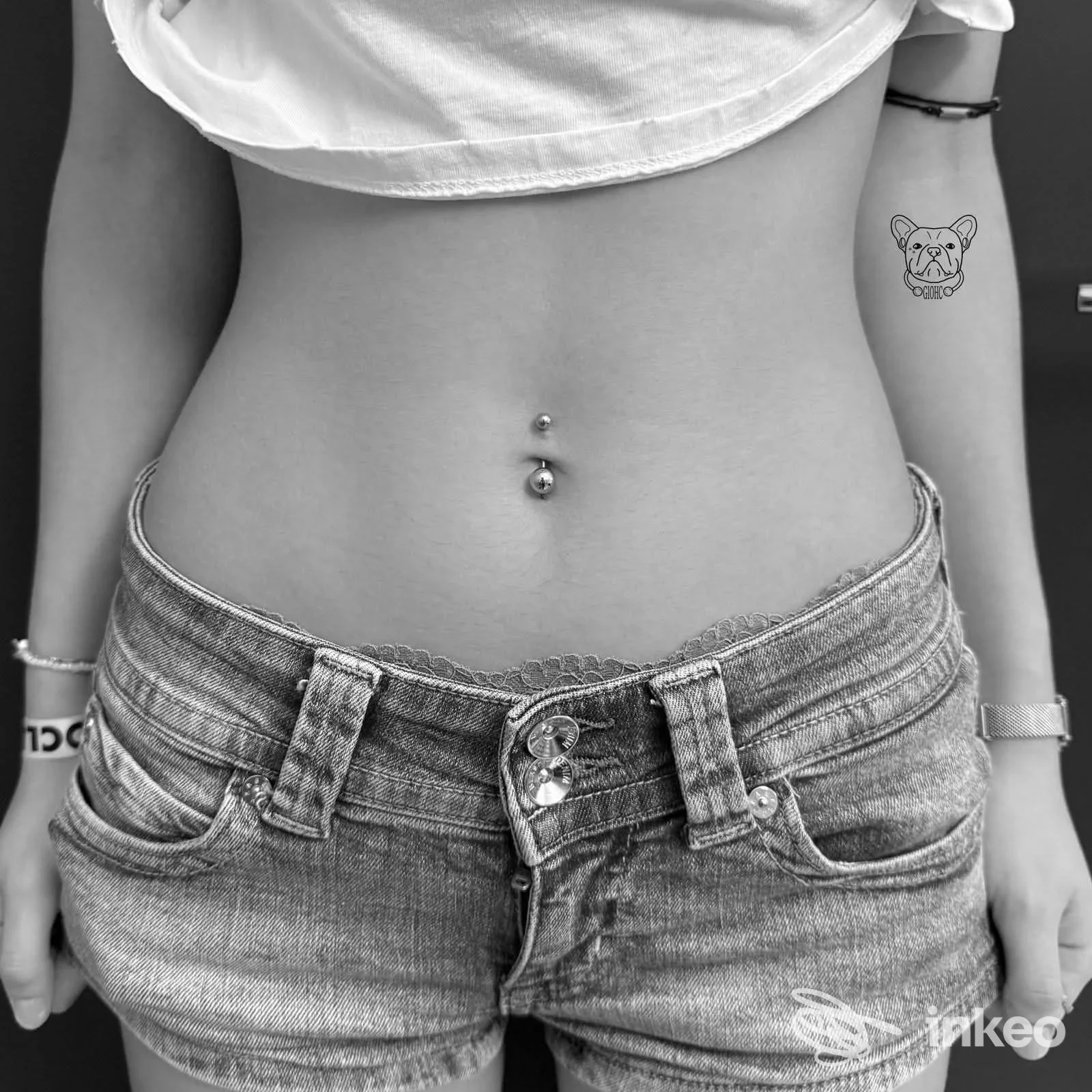 Una modella mostra addome nudo con piercing all’ombelico e una piccola tatuatura di cane sul braccio destro, indossa jeans corti e una maglietta tagliata.