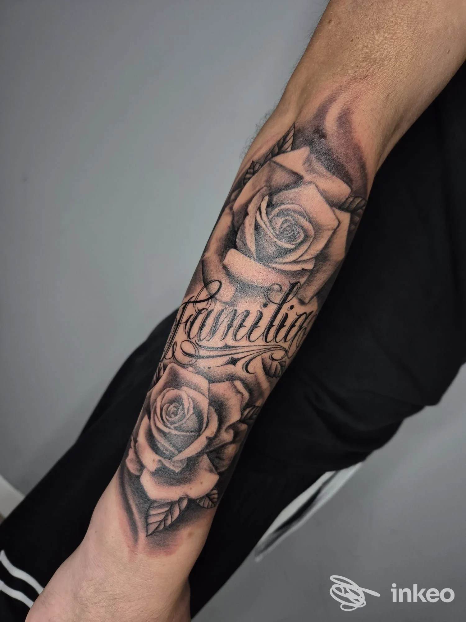 Braccio con tatuaggio visibile raffigurante rose e scritte, senza volto visibile nè testo promozionale; si vede una parte dell’avambraccio e una mano sullo sfondo, in bianco e nero-grigiastro.