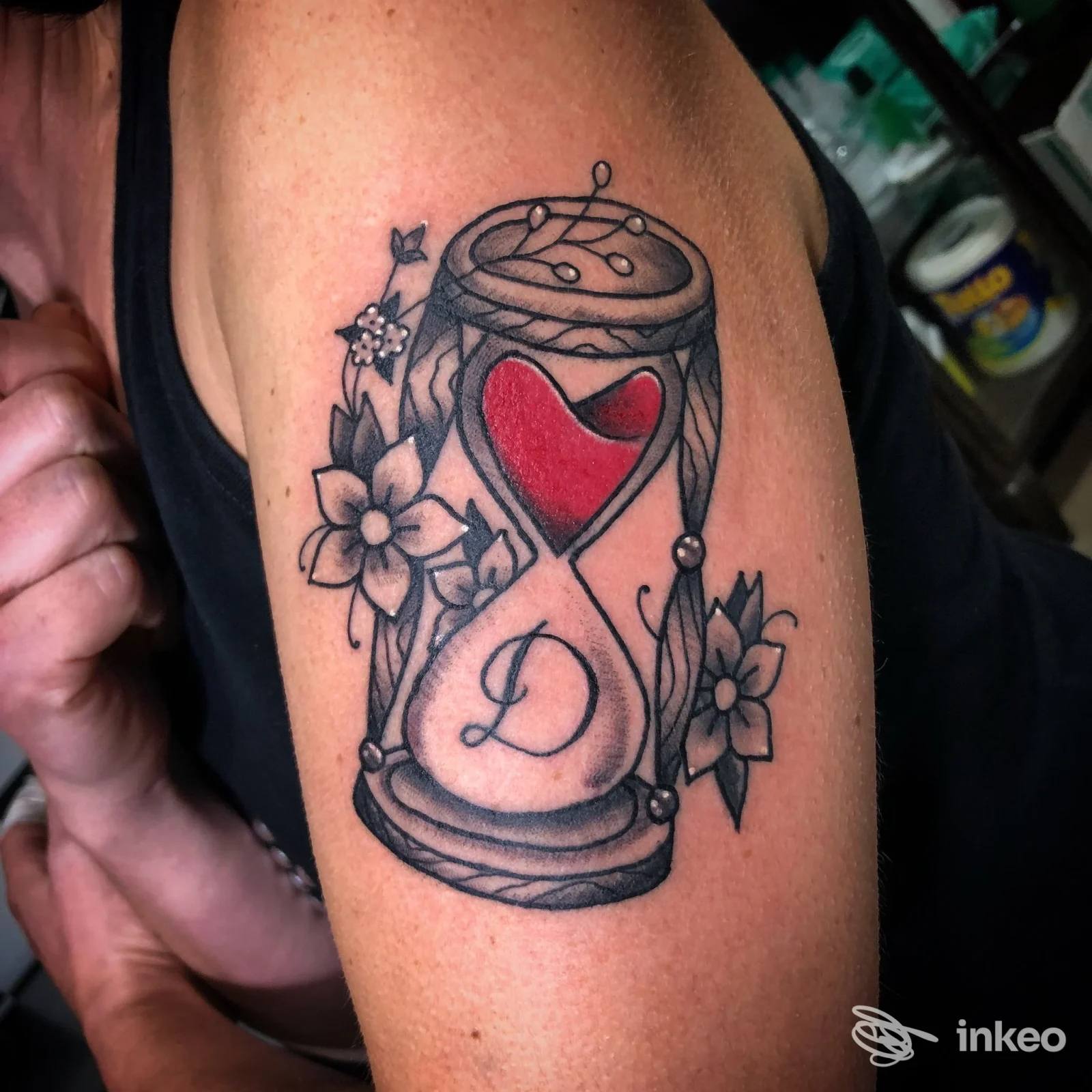 Tatuaggio su braccio raffigurante una clessidra con cuore rosso e fiori, contorni in nero e dettagli di shading.