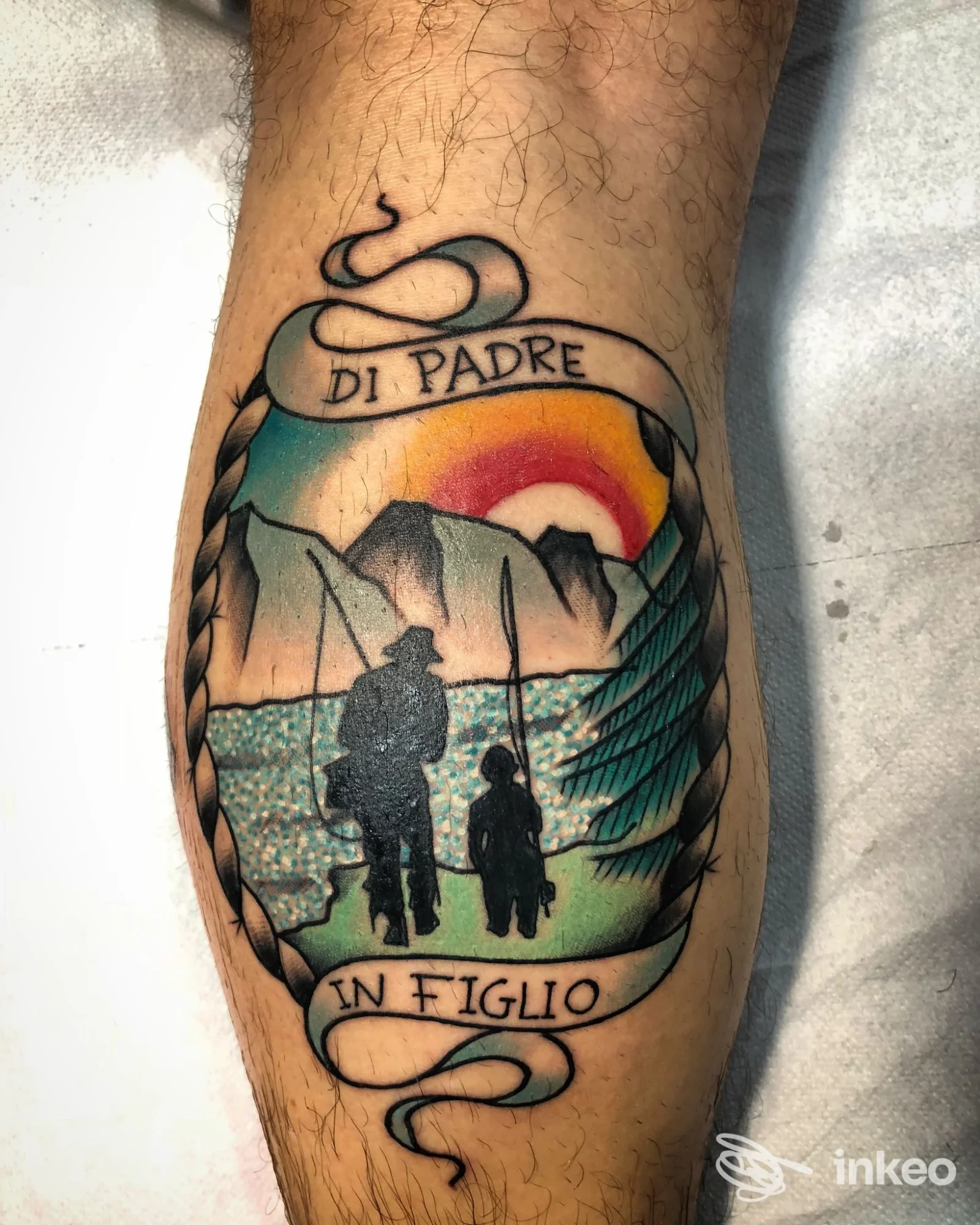 Tattoo a colori su polpaccio raffigurante due figure al tramonto tra montagne e mare, con cornice a nastro intrecciato. Foto vicino che mette in risalto i dettagli del disegno.