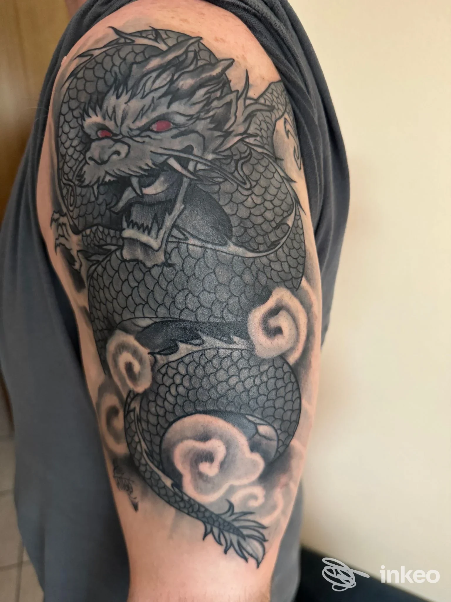 Tatuaggio drago nero/grigio su braccio, con scaglie definite, occhi rossi e nuvoletta di fumo, occupa la parte superiore del braccio e la spalla.