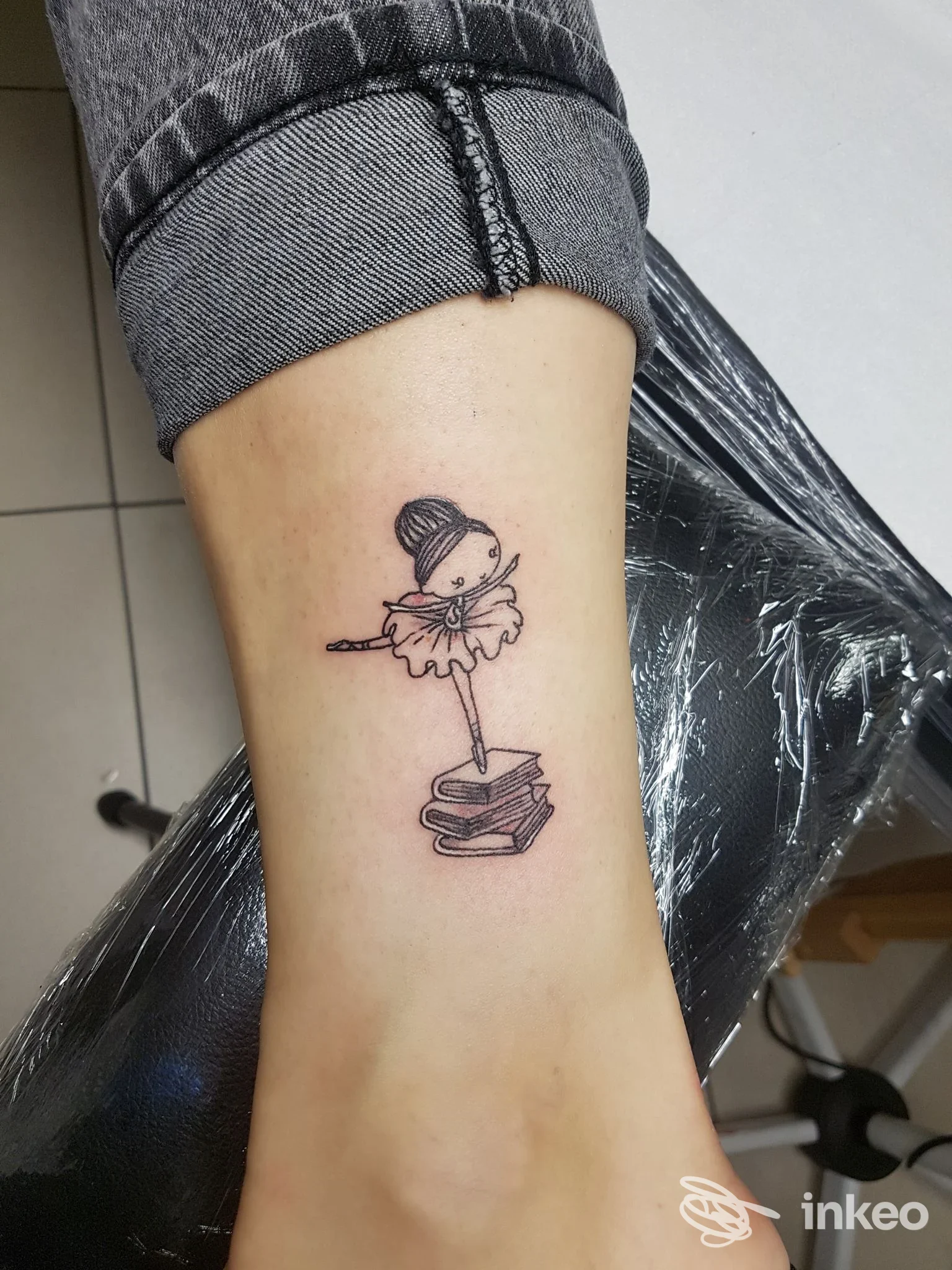 Avambraccio con piccolo tatuaggio: una ballerina appoggiata su una pila di libri, disegno a linee fini, senza volto umano visibile.