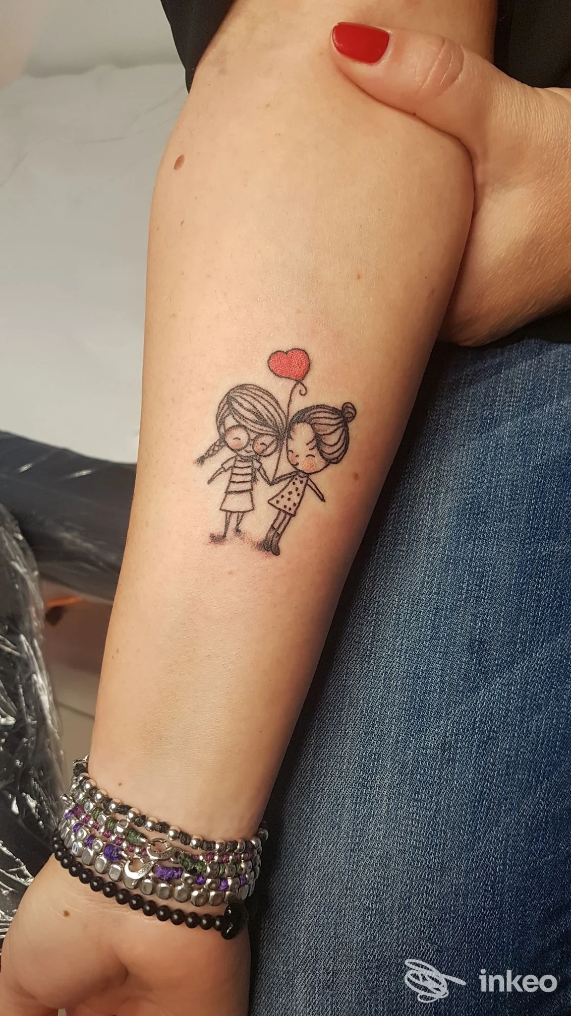 Tattoo su avambraccio raffigurante due bambini che tengono un palloncino a forma di cuore, tratto in nero con dettagli rossi, visibile sull’avambraccio della persona.