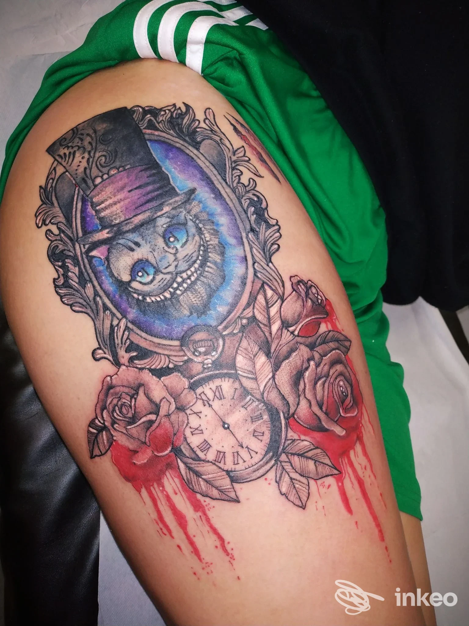 Tatuaggio sul braccio raffigurante Cheshire Cat entro cornice decorativa, orologio da tasca e rose, con gocciolamenti rossi.