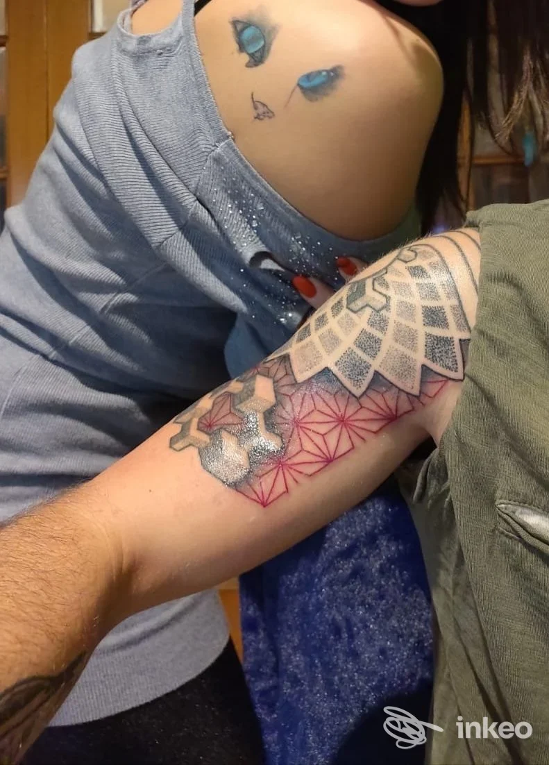 Avambraccio visibile con un tattoo geometrico a rete di tonalità rosa, contorni neri, in primo piano; due persone inquadrate, nessun volto interamente visibile.