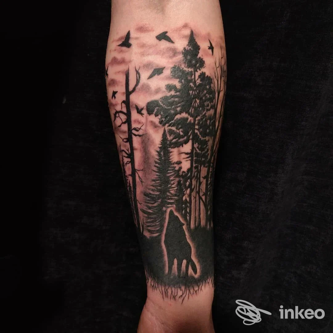 Tatuaggio sul braccio raffigurante un lupo accanto a una foresta con alberi alti, volti uccelli in cielo rosa.