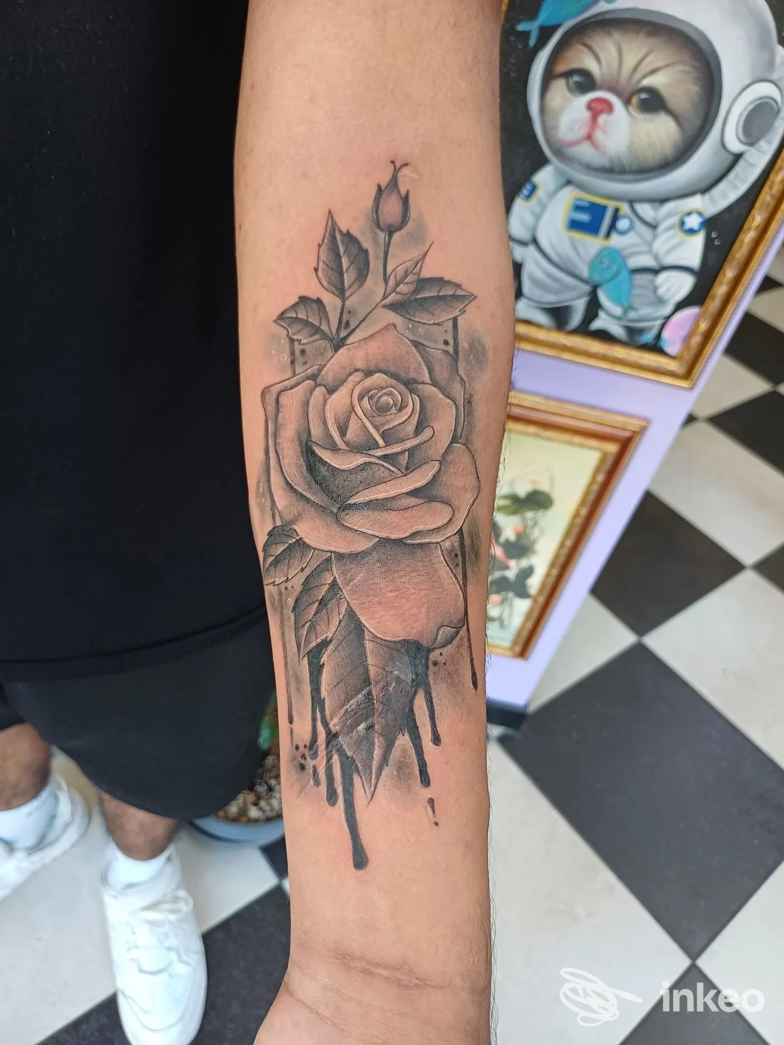 Tatuaggio su avambraccio raffigurante rosa con sfumature grigie e dettagli realistici, visibile davanti a pavimento a scacchi e cornici di quadri sullo sfondo.