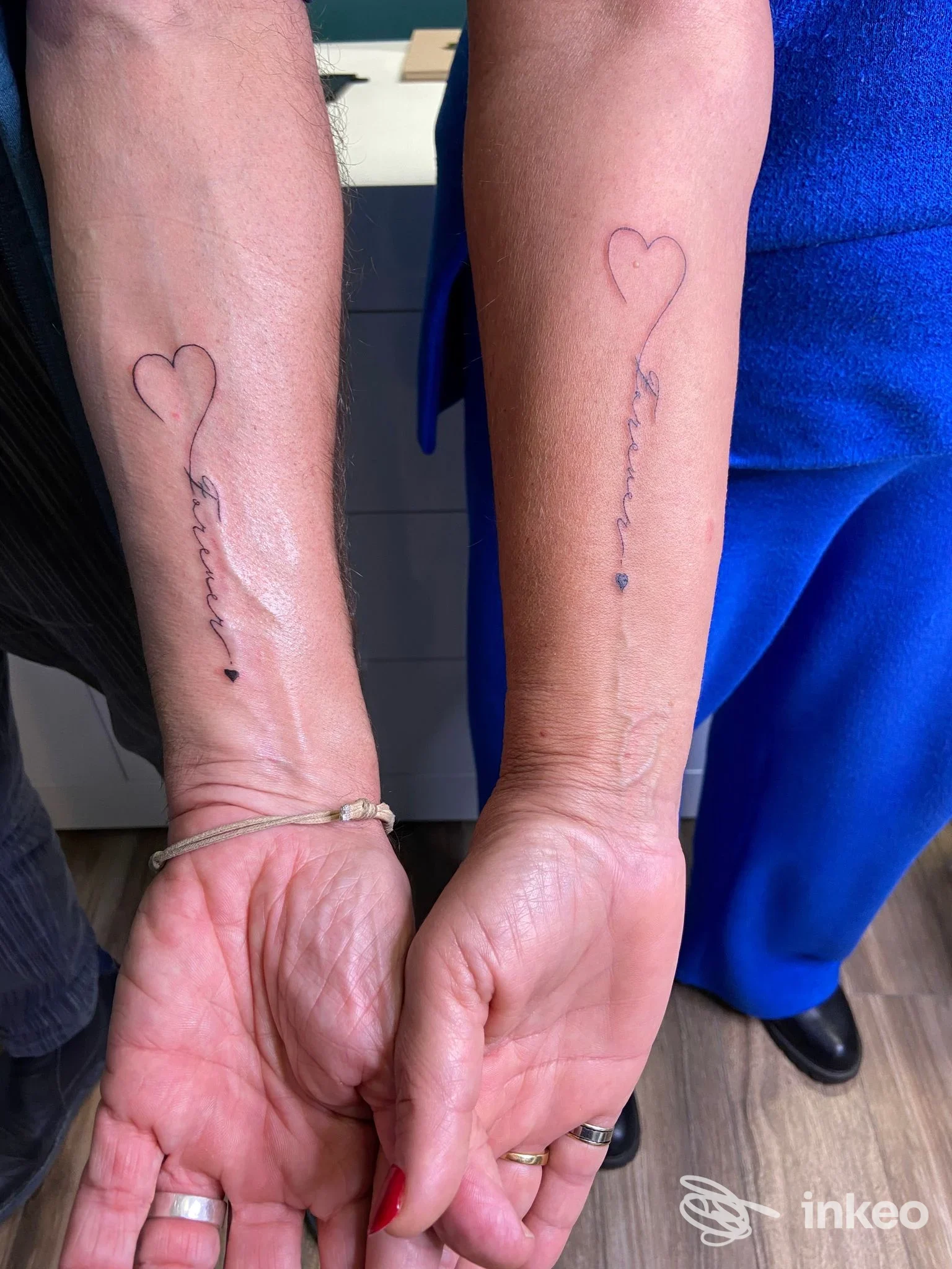 Due avambracci con tatuaggi coordinati a Budrio realizzati da DermaUp: cuore contorno e linea script sottile sul lato interno dell'avambraccio.