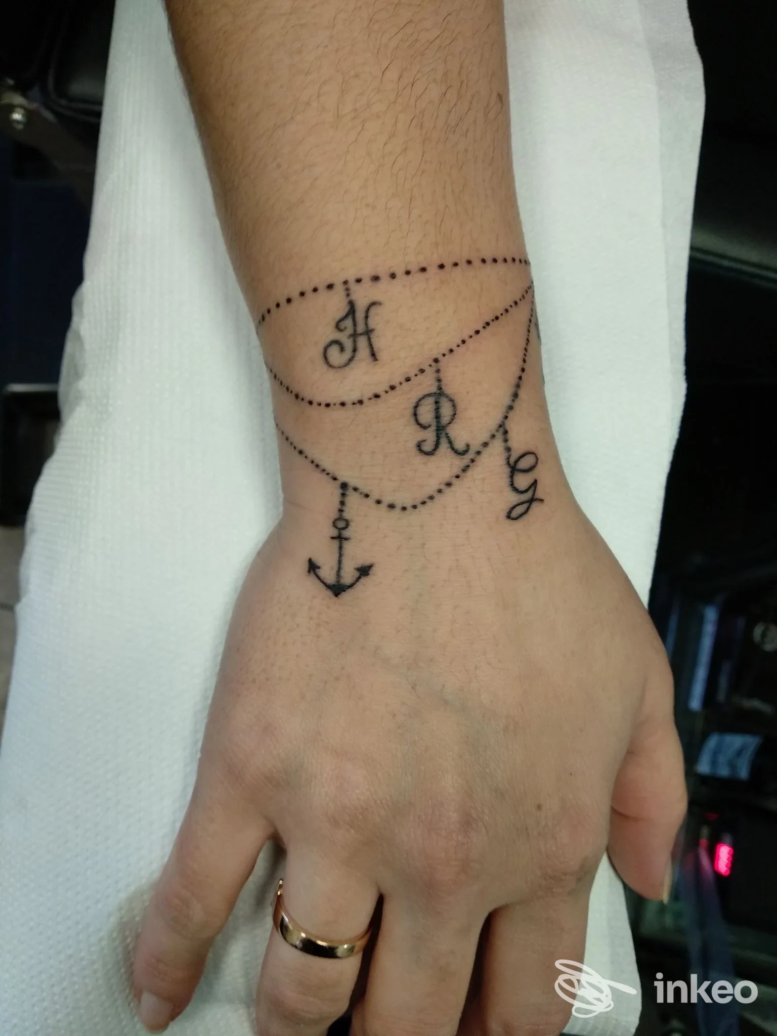 Tatuaggio nero su polso e dorso della mano con catene punteggiate, iniziali 'H R G' e una piccola ancora – The Mood Tattoo, Vigolzone.