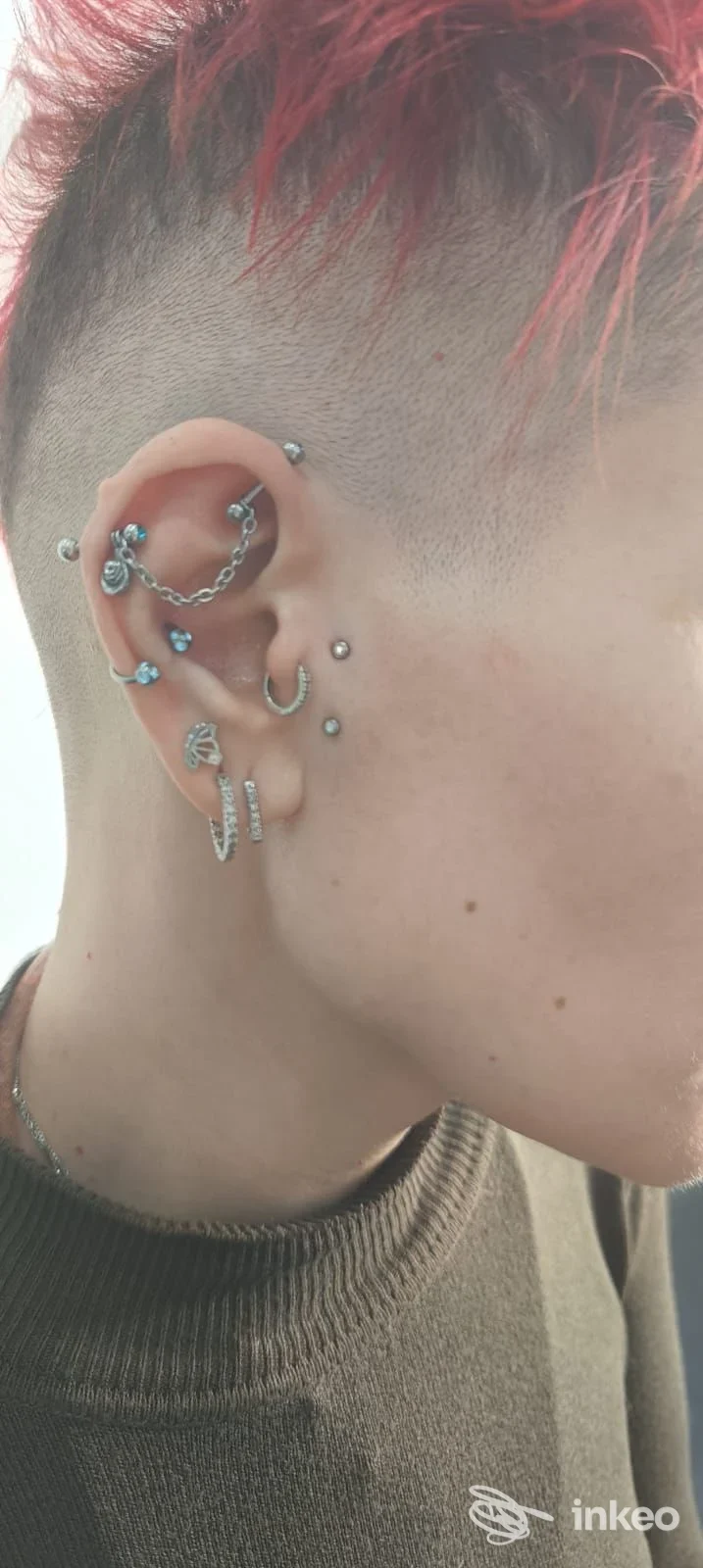 Primo piano laterale di una persona con capelli rasati e colorati che mostra molteplici piercing all'orecchio (catena, cerchi e microgemme). Foto per More Piercing e PMU, Collecchio.