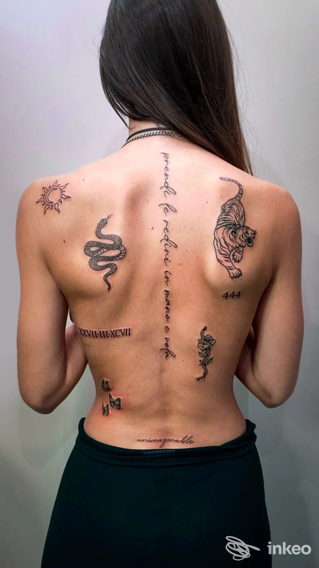 Schiena di persona da dietro con vari tatuaggi: serpente, scritte verticali e altri motivi, senza elementi di interfaccia sociale visibili, ritraente la pelle tatuata.