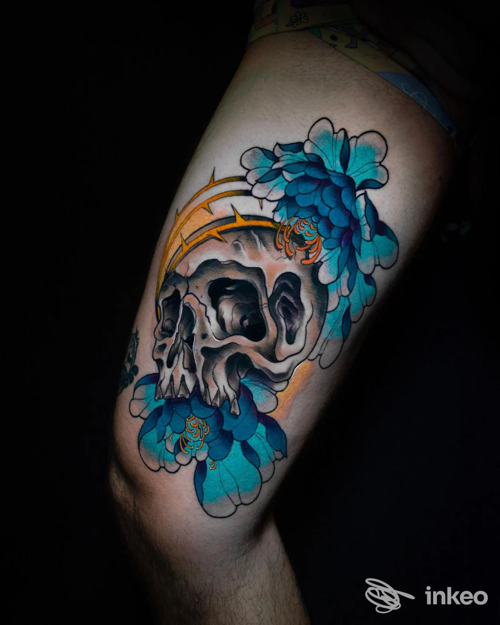 Tatuaggio su coscia raffigurante un teschio realistico avvolto da grandi fiori blu e dettagli arancioni, foto per Crazy Fellas, Bologna.