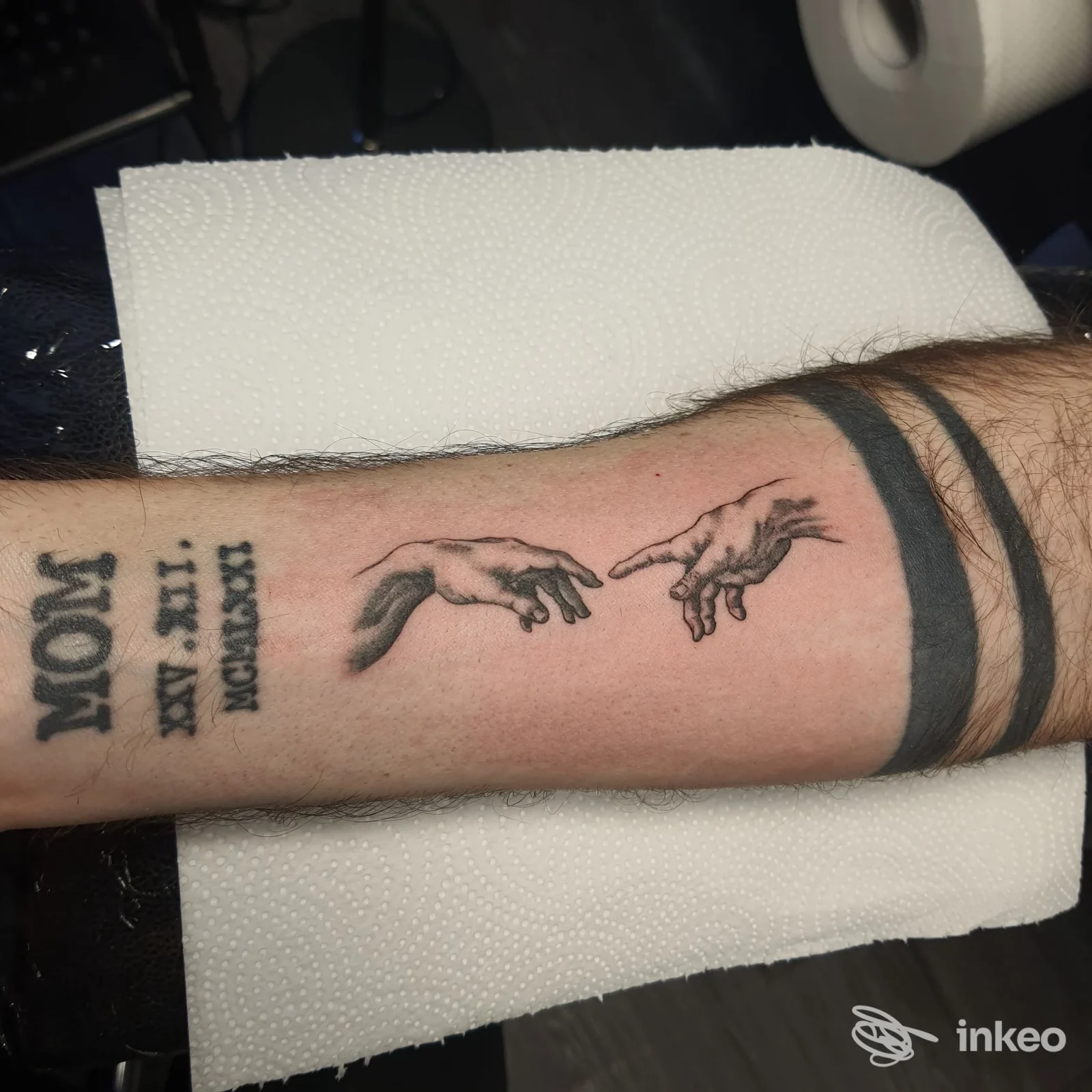 Primo piano dell'avambraccio con tatuaggi: mani ispirate alla Creazione, fasce nere e scritte. Art Design Ink, Cervia.