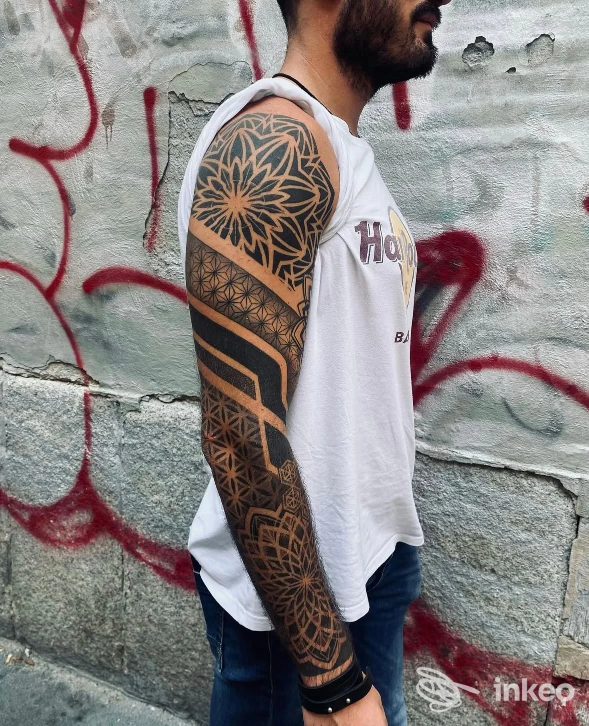 Tonico tatuaggio geometrico nero su braccio sinistro, visibile in primo piano, sfondo con graffiti; persona reale presente.