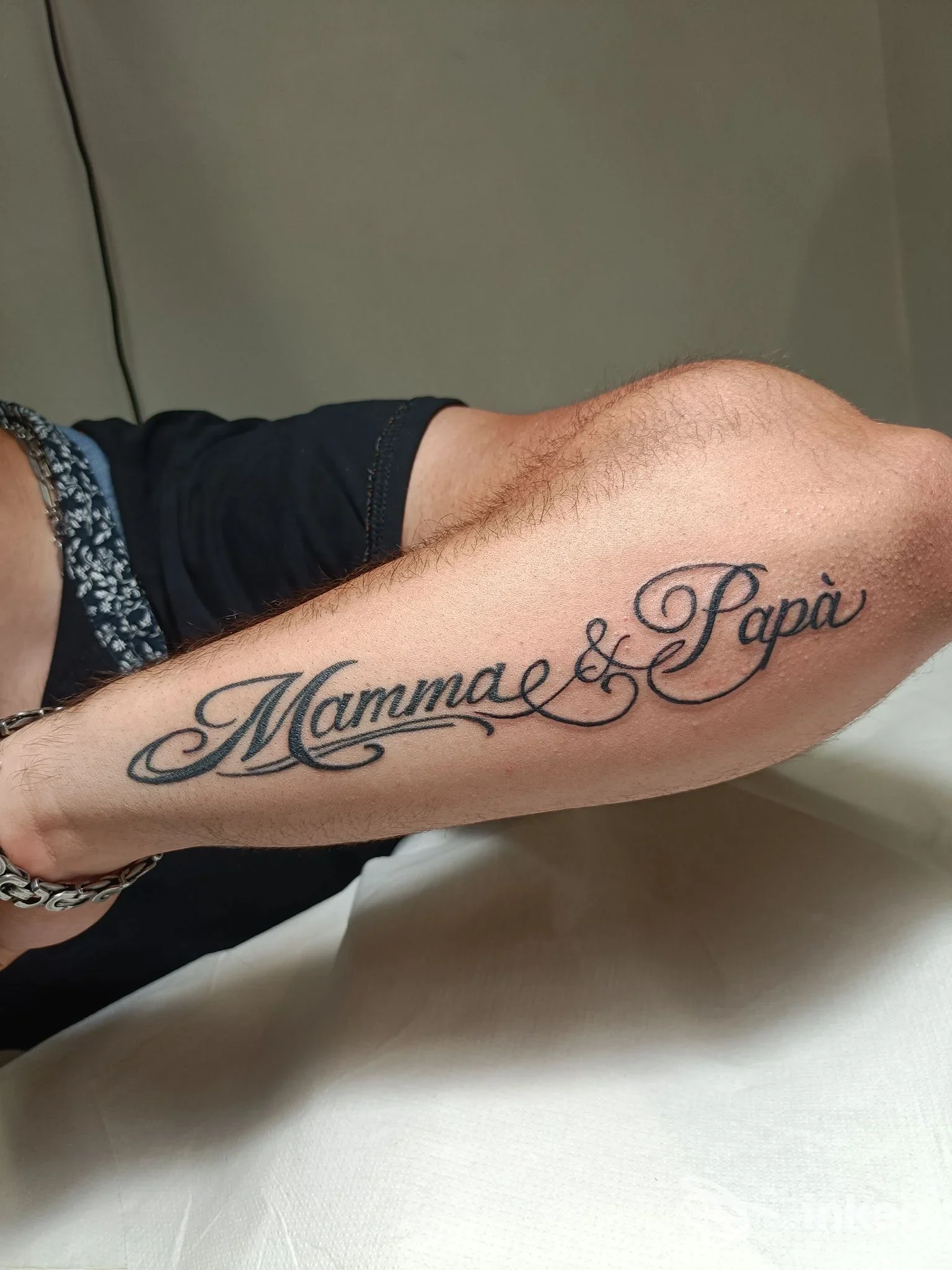 Tatuaggio scriptato sul antebraccio sinistro con la scritta 'Mamma & Papà' collegata da un motivo decorativo; pelle nuda visibile.