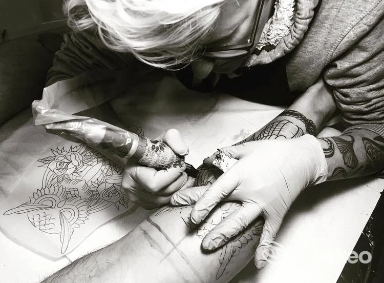 Tatuatore con guanti sta inchiostrando un braccio: la macchina da tatuaggio è infilata nella pelle e si vedono schizzi sul tavolo.