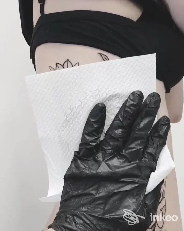 Dettaglio di un tatuaggio floreale sul fianco di una persona, con guanto nero e carta assorbente, Novytattoo Handmade Tattoo Studio, Carpi.