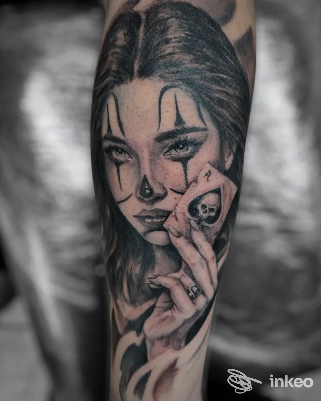 Tatuaggio realistico in bianco e nero su avambraccio: volto femminile trucco clown e mano con carta teschio — Infamilia Tattoo, Sassuolo.