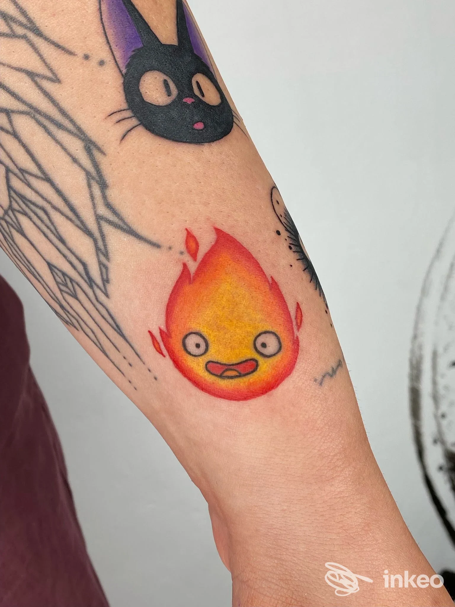 Tattoo sul braccio: una piccola fiametta arancione con faccina, visibile accanto ad altri elementi del braccio.