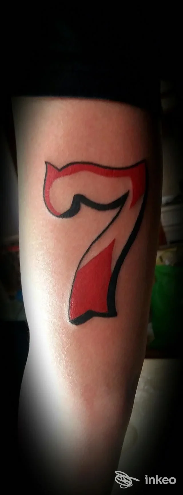 Braccio con tatuaggio a forma di numero sette, contorno nero e riempimento rosso, visibile sulla pelle.