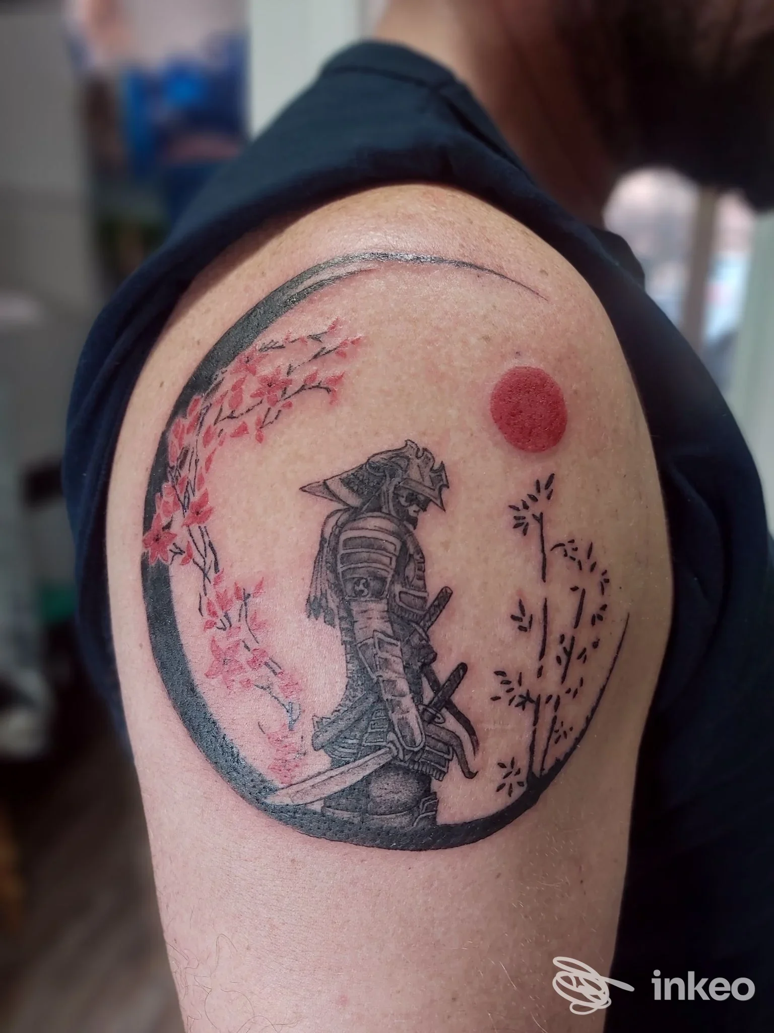 Tatuaggio a tema samurai sulla spalla, eseguito da La Tatueria della Sha a Villa Minozzo, dettaglio con fiori e disco rosso.