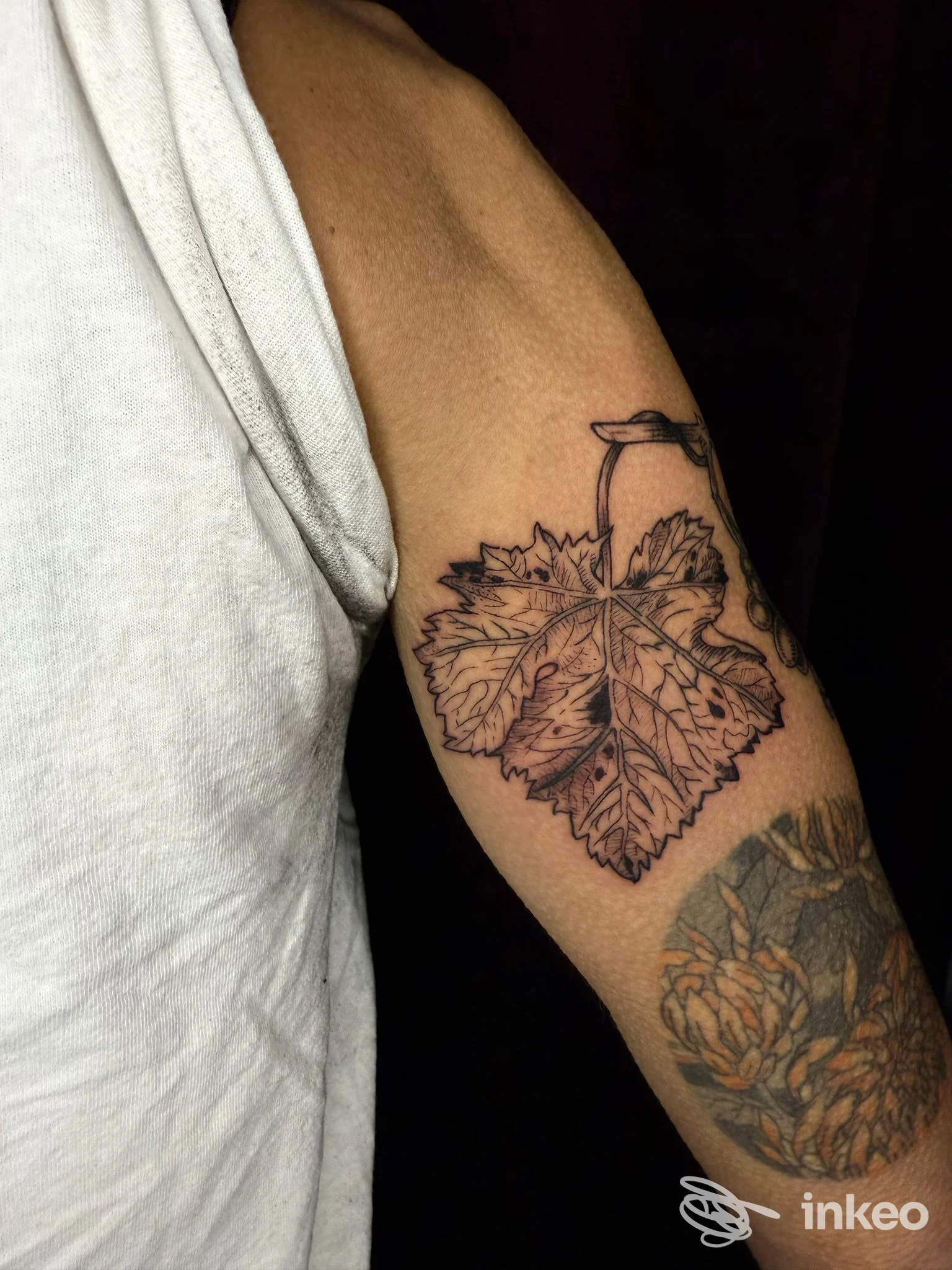 Braccio sinistro con due tattoo: una grande foglia decorativa sul bicipite e un secondo motivo floreale parzialmente visibile sull’avambraccio, indossando una maglietta bianca.