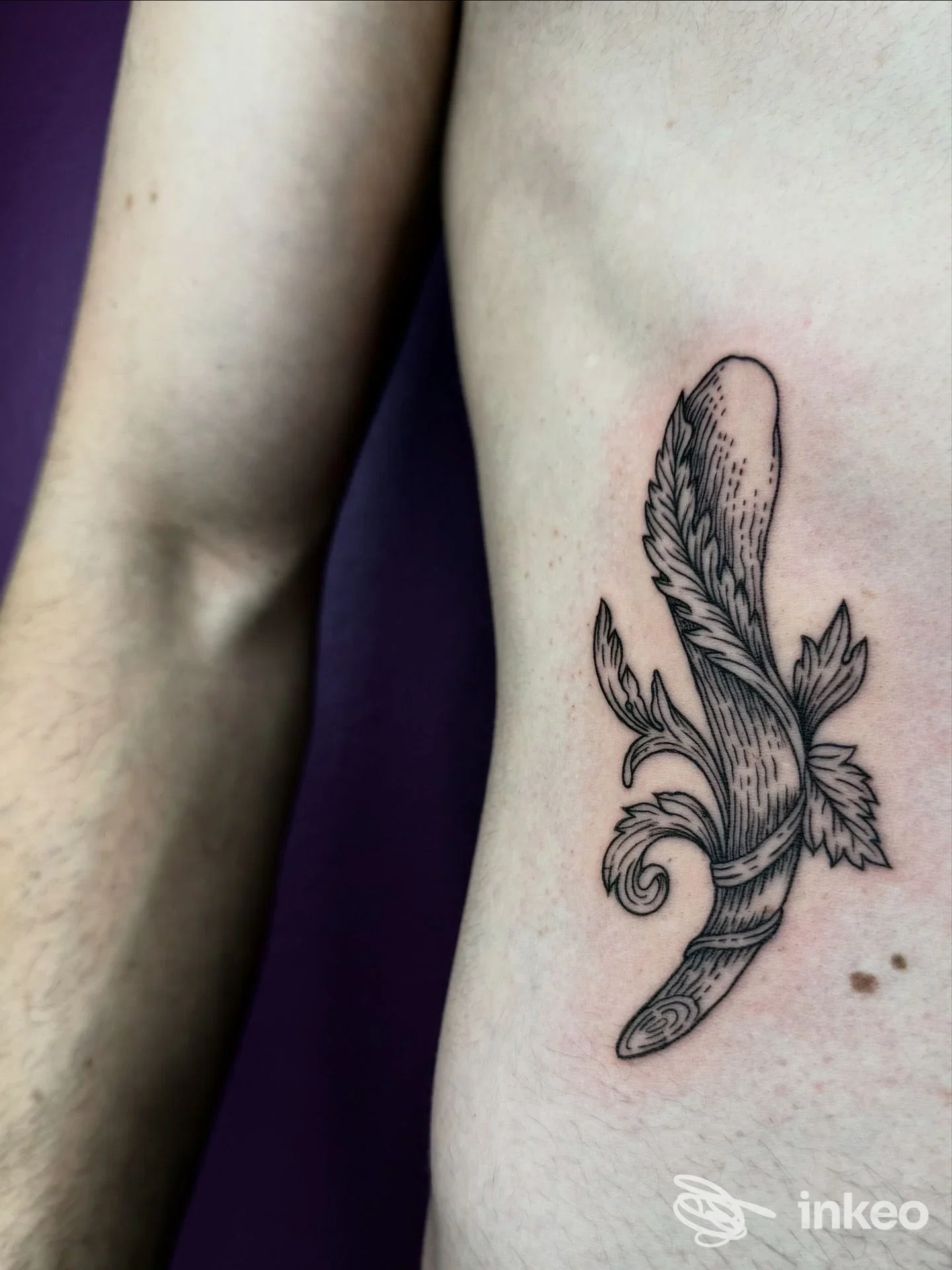 Tattoo su pelle umana raffigurante una piuma stilizzata con dettagli in linework nero, posizionato sul fianco/avambraccio.