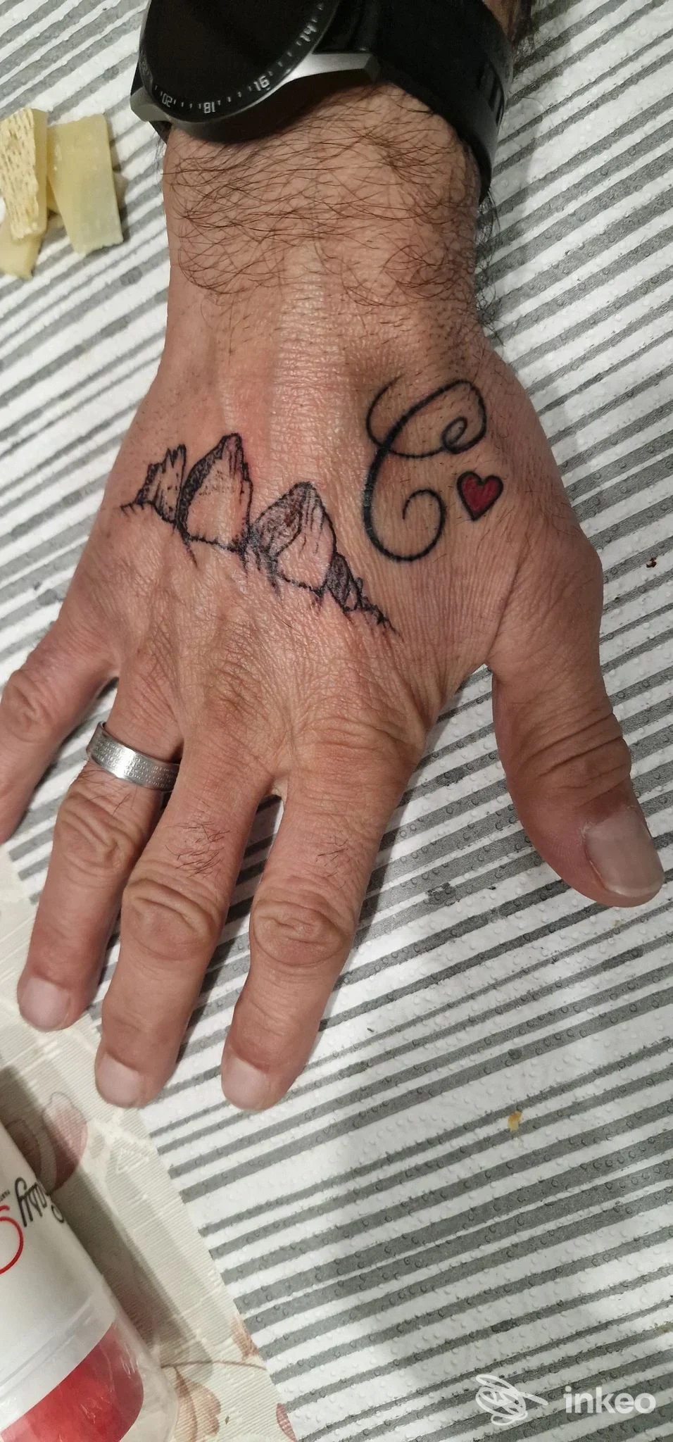 Dorso della mano tatuato con una linea di montagne, lettera C calligrafica e piccolo cuore. Ink On Show Tattoo e Piercing, Rimini.