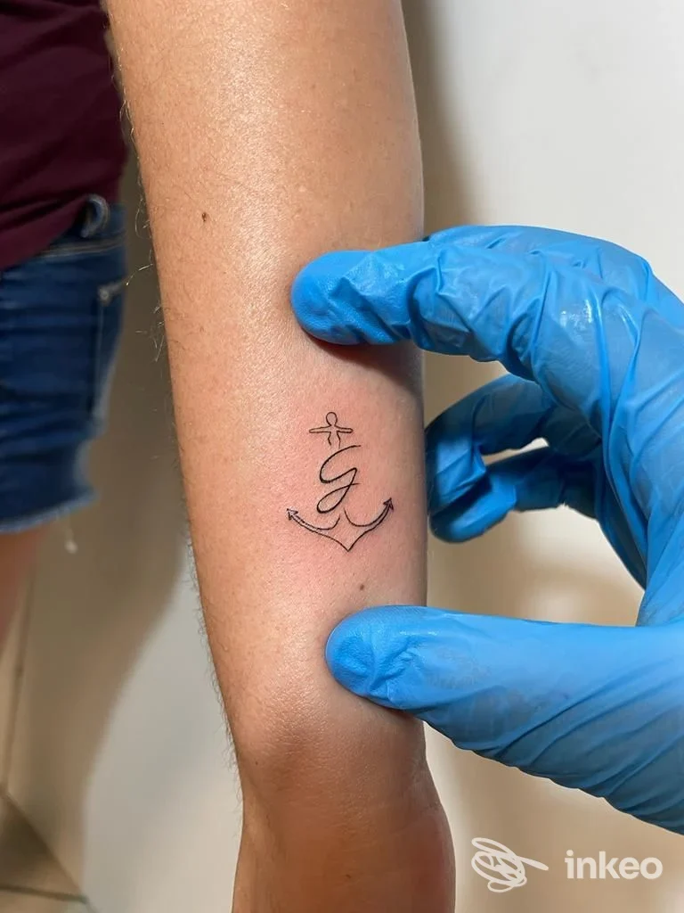 Avambraccio con piccolo tatuaggio minimal a forma di ancora e lettera, foto da Tropical Tattoo a Guastalla, mani con guanti blu dello tatuatore.