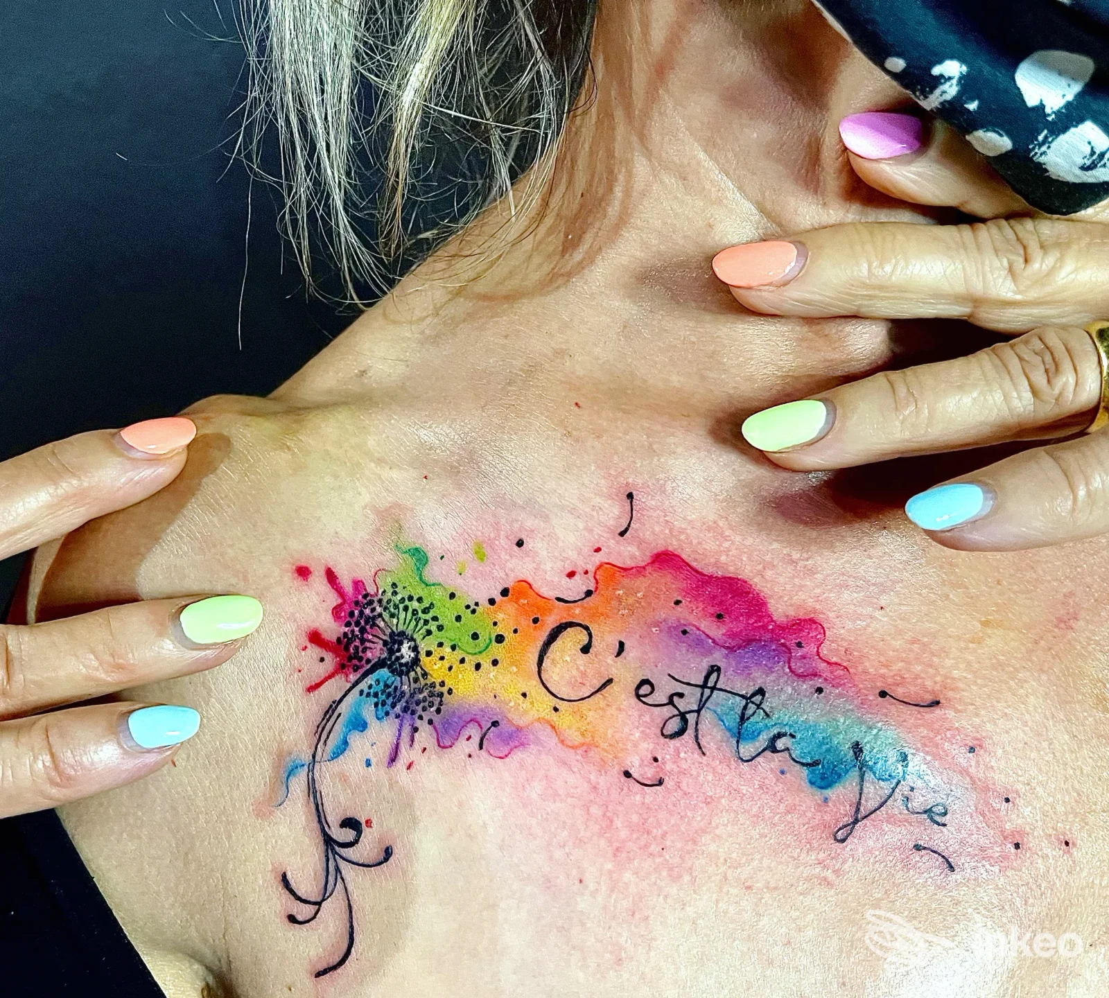 Tattoo ad acquerello colorato sulla spalla sinistra di una cliente, con riflessi rosa, azzurro e verde, bordi scuri e linee fluide; mani con unghie colorate tengono la cute ben visibile.