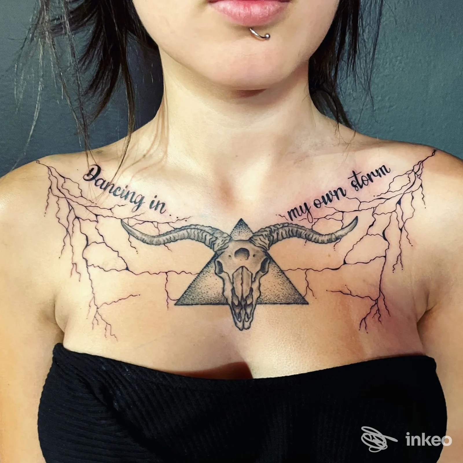 Tattoo sul décolleté raffigurante un cranio di capra inserito in un triangolo, con fulmini ai lati e la scritta Dancing in my own storm lungo la linea delle clavicole.