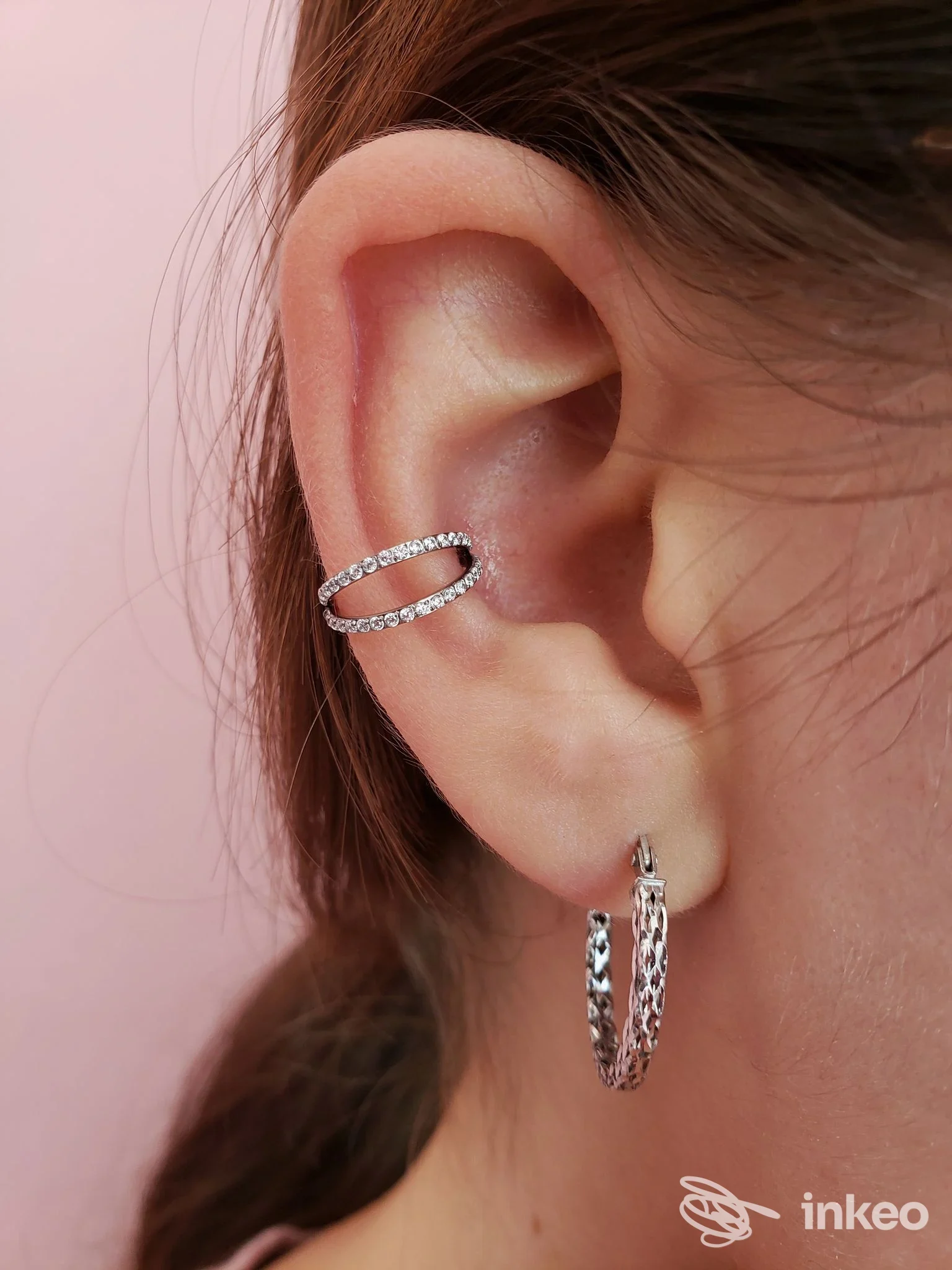 Primo piano dellorecchio sinistro di una persona, mostrante due piercing: un cerchio a ferro al lobo e una clip/ear cuff all’alto dell’orecchio, con sfondo rosa chiaro.
