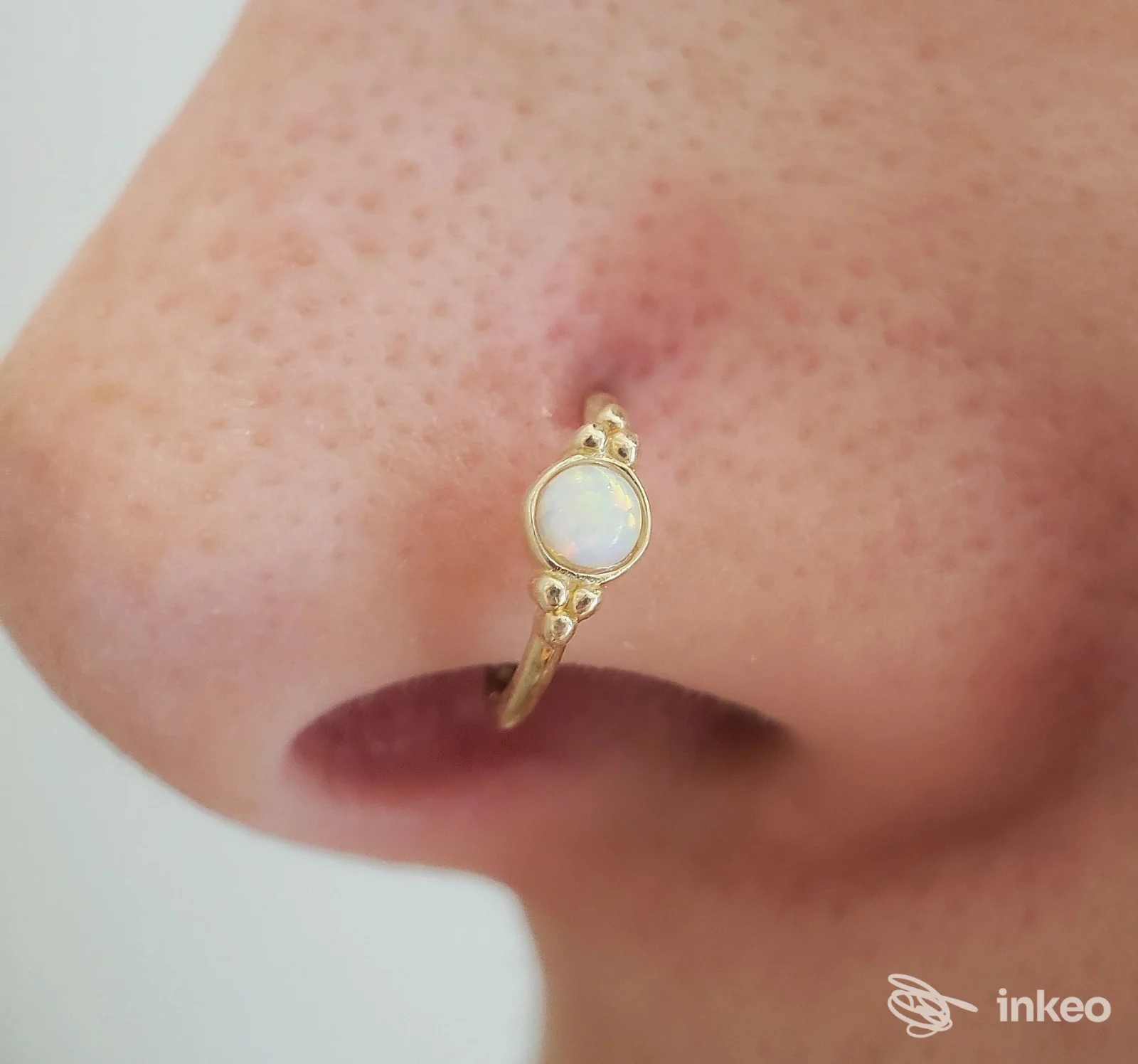 Anello piercing dorato con pietra opale posizionato sul naso di una persona, in primo piano vicino all’estremità del naso, con sfondo neutro.