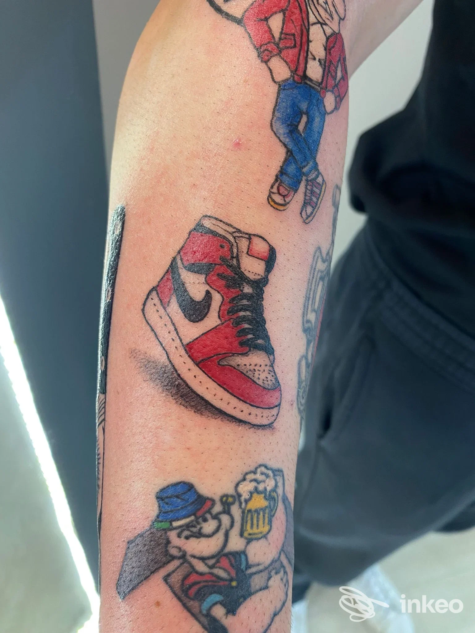 Primo piano di avambraccio con tatuaggi illustrativi: una sneaker e un personaggio con una pinta, Noa Noa Beach Tattoo, Lido di Savio.