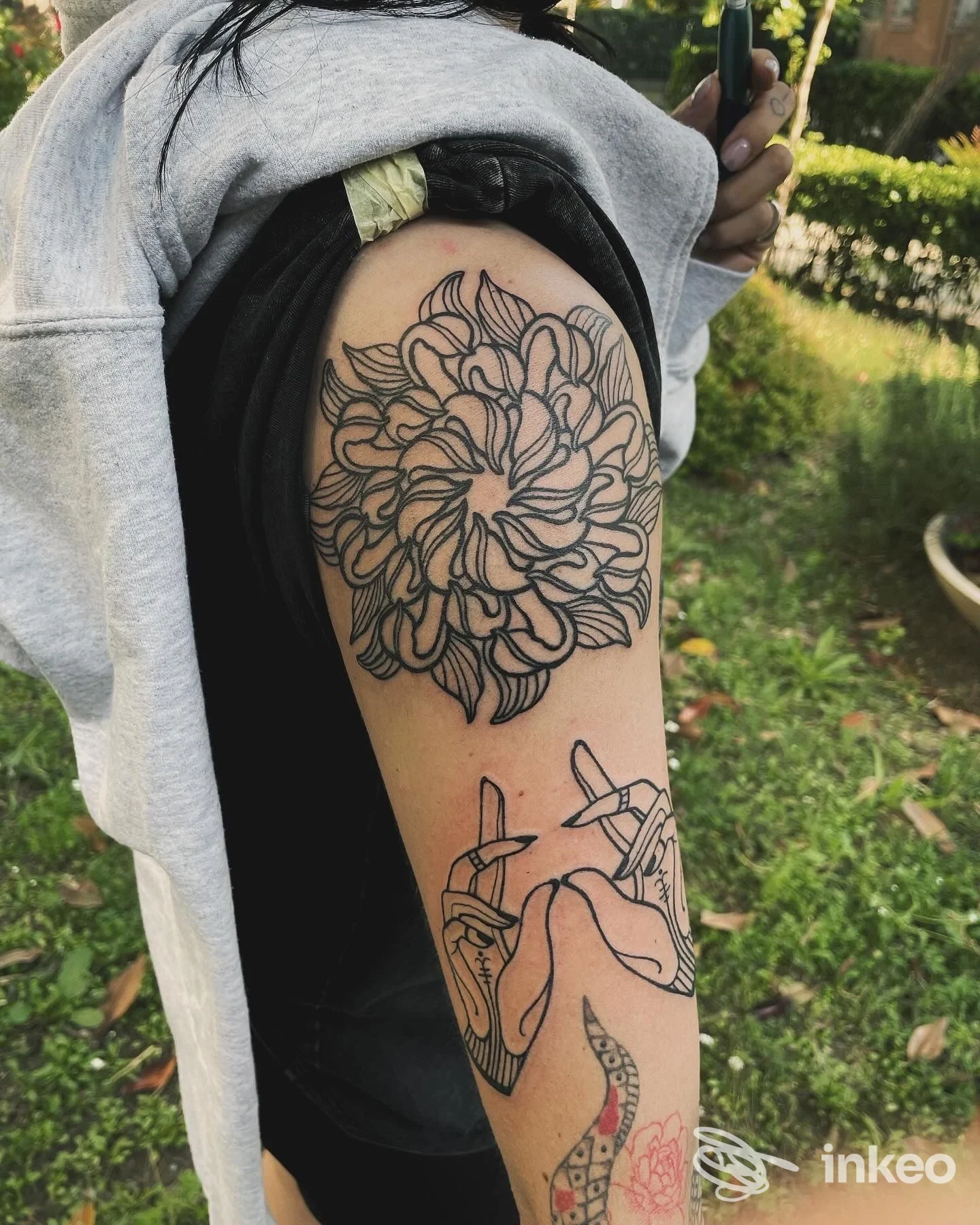 Braccio con due tatuaggi: grande fiore stilizzato sulla spalla e mani incrociate in linework. Foto fornita da Magma Tattoo Studio, Riccione.