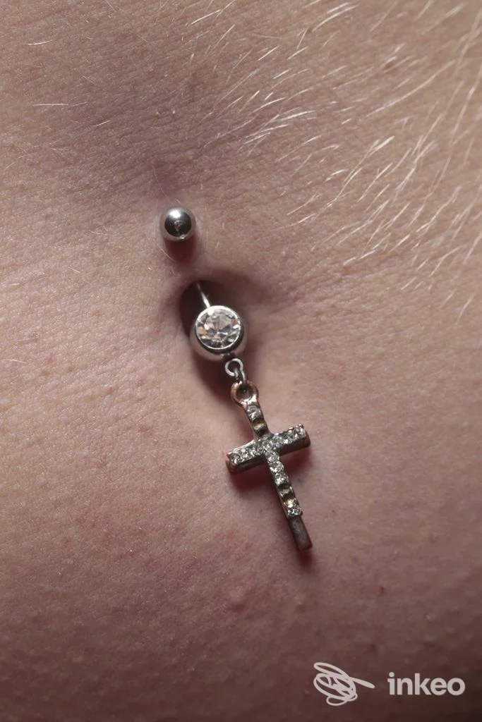 Piercing ombelicale con pendente a croce in metallo lucido, dettaglio sulla pelle. Nonsolopiercing Studio, Udine.
