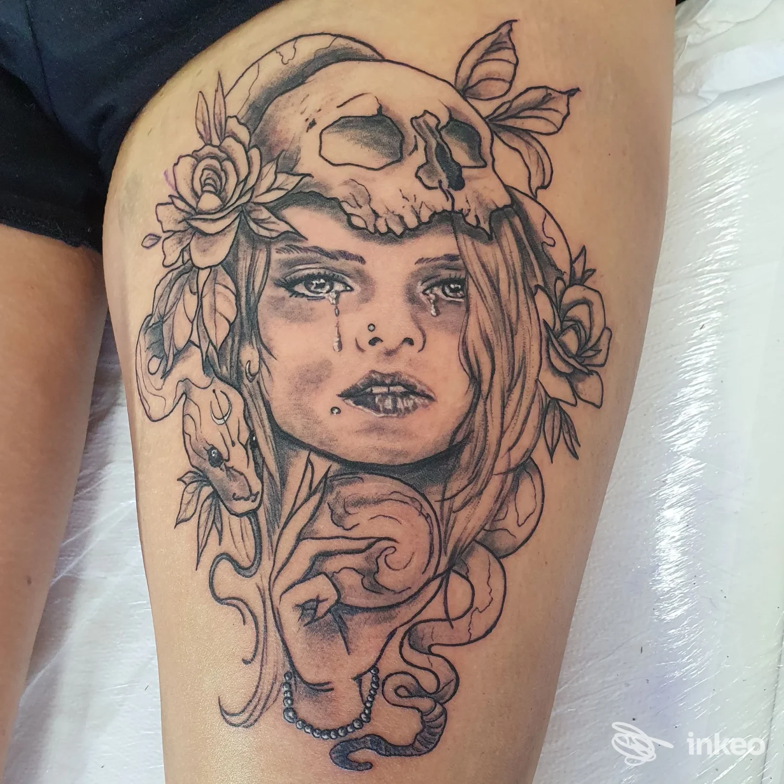 Primo piano di un tatuaggio in bianco e nero su coscia: volto femminile con cranio, fiori e serpente. Wanderlusttattoo, Buia.