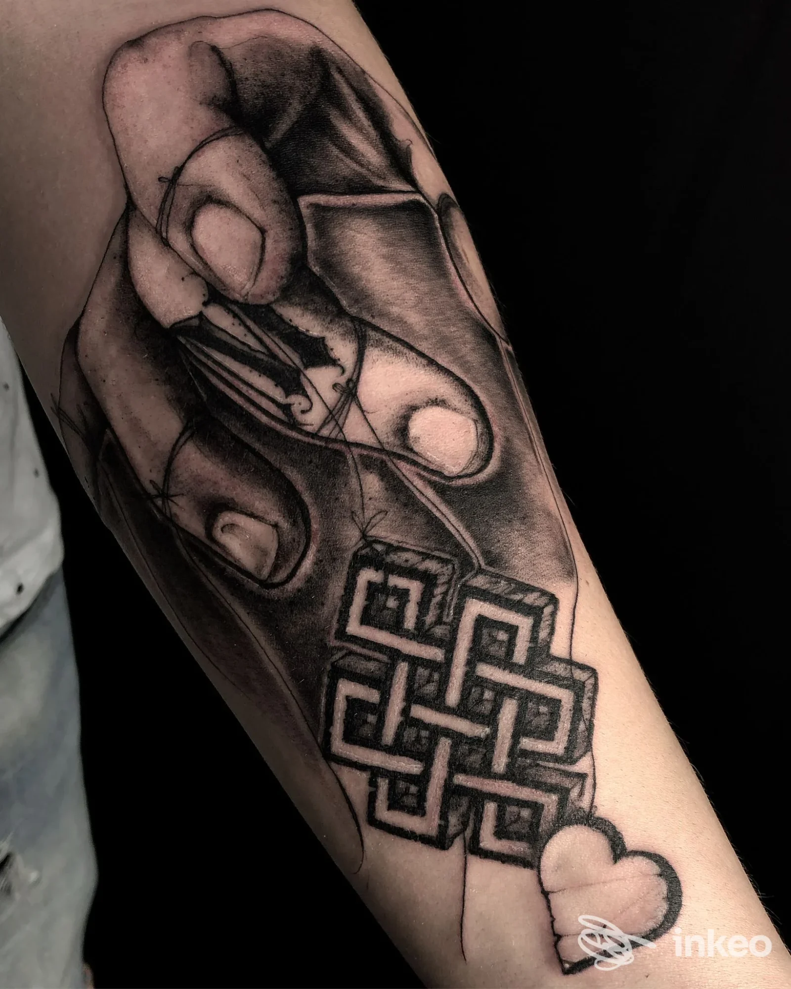 Tatuaggio in bianco e nero sull'avambraccio raffigurante una mano che tiene un pendente con nodo geometrico e un cuore, realizzato da SGB Tatuaggi e Piercing a Gorizia.