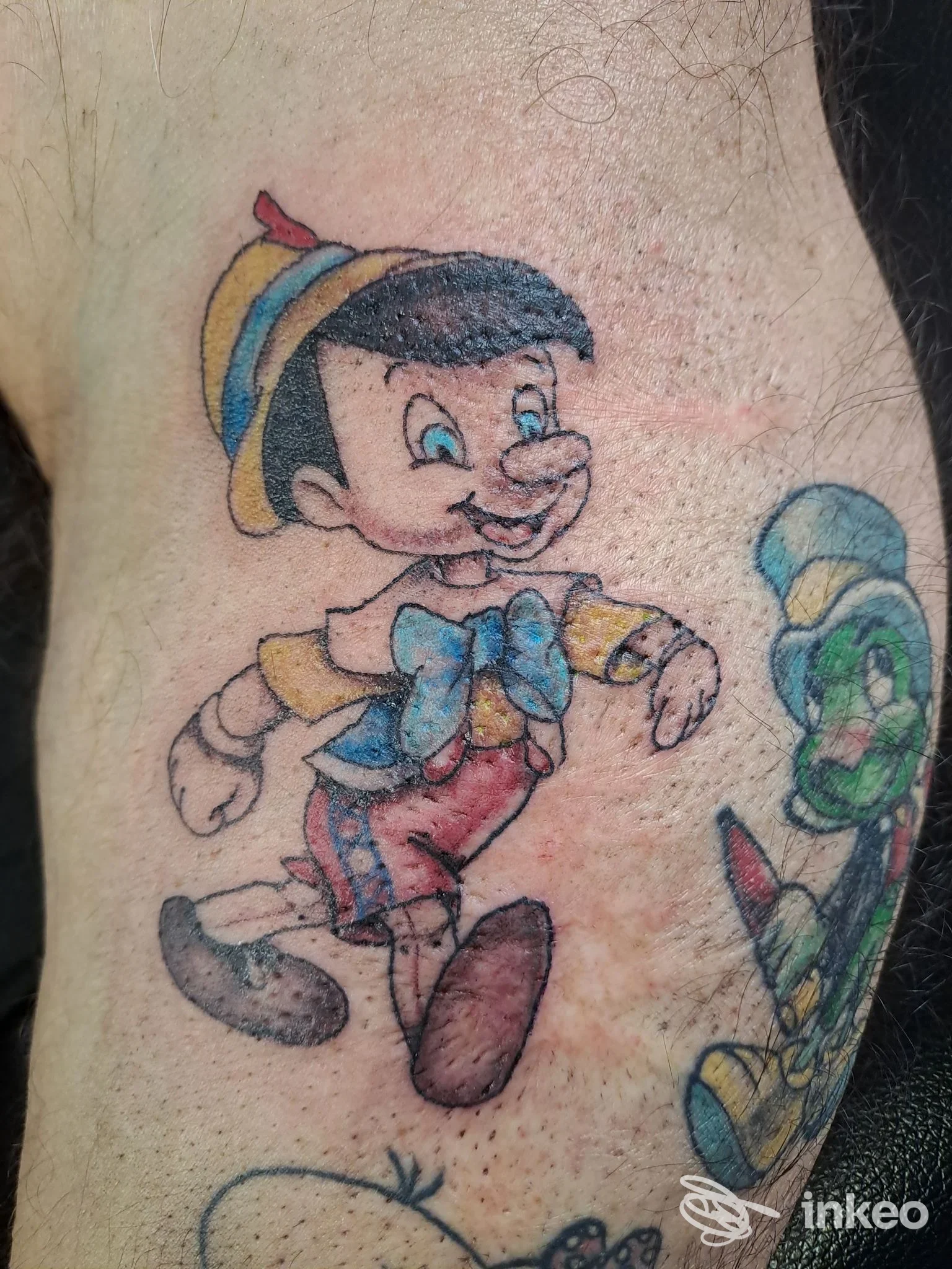 Tattoo colorato su pelle umana raffigurante un personaggio stile Pinocchio con cappello e pantaloni rossi, in posizione dinamica sulla pelle del braccio/sopra spalla.