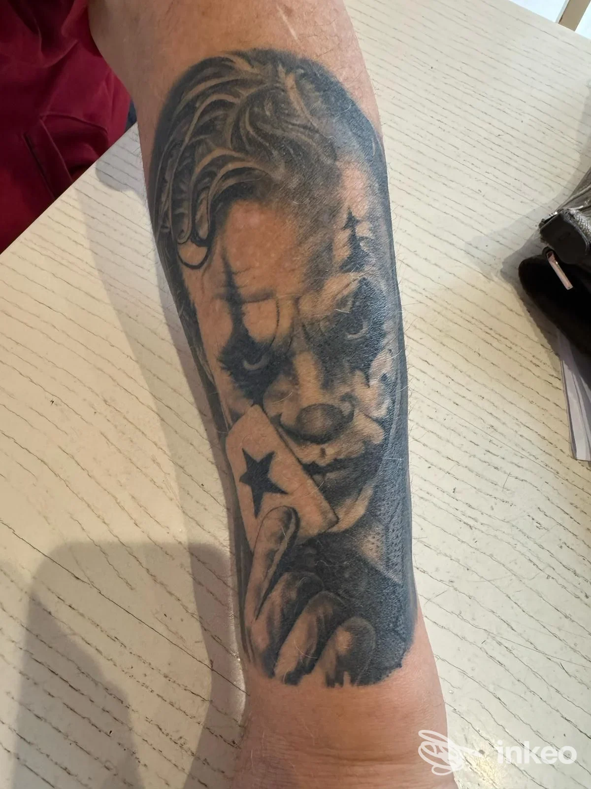 Tatuaggio in bianco e nero sull'avambraccio con volto clown realistico che tiene una carta — Peto Tattoo, Sacile.