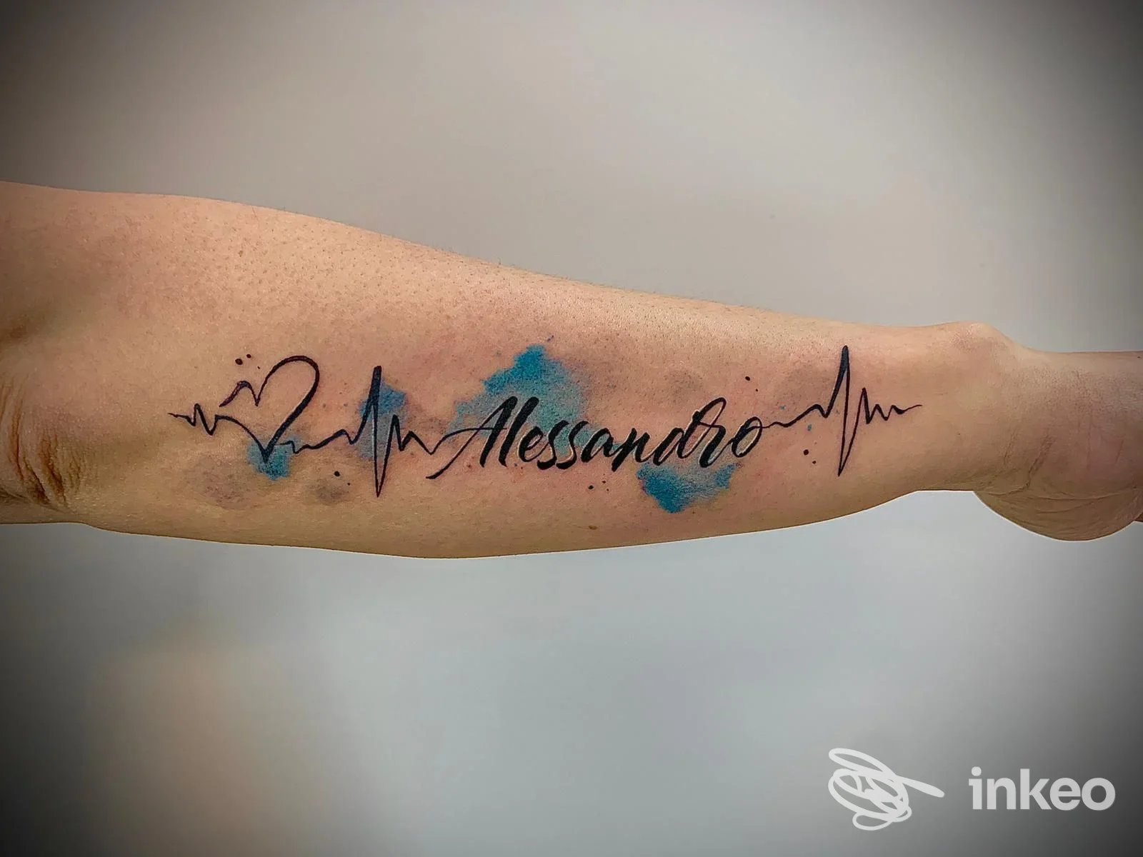 Tatuaggio con nome 'Alessandro' e battito cardiaco su avambraccio, effetto acquerello blu. Realizzato da Inkheart Tattoo Studio, Staranzano.